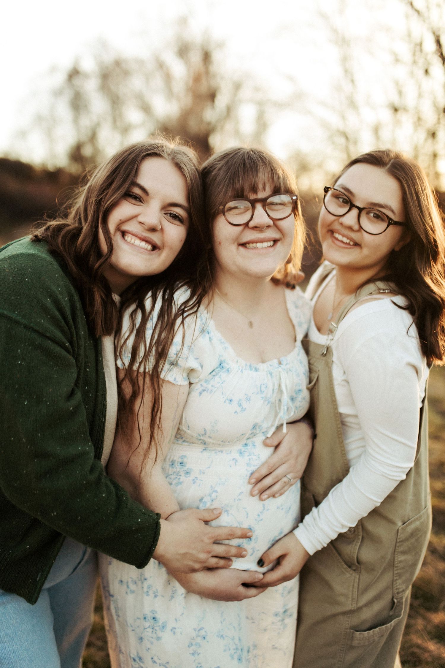 Sisters all standing together closely and joyfully for family photos, middle sister holding her pregnant belly smiling brightly