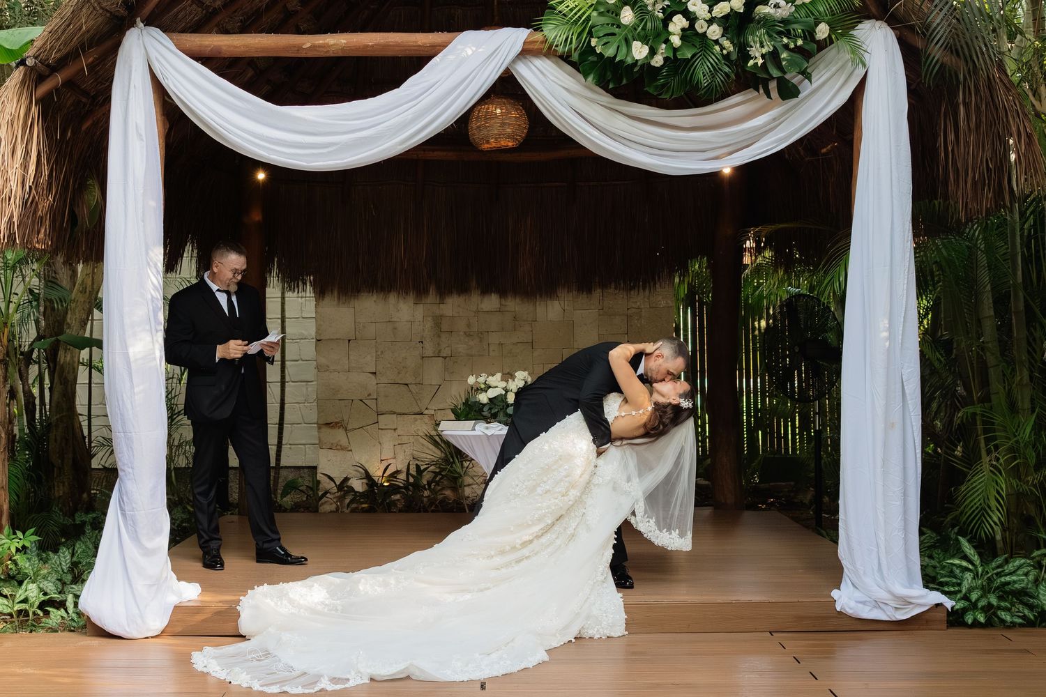 Dramatic dip pose under white draped wedding arch decorated with greenery and flowers.