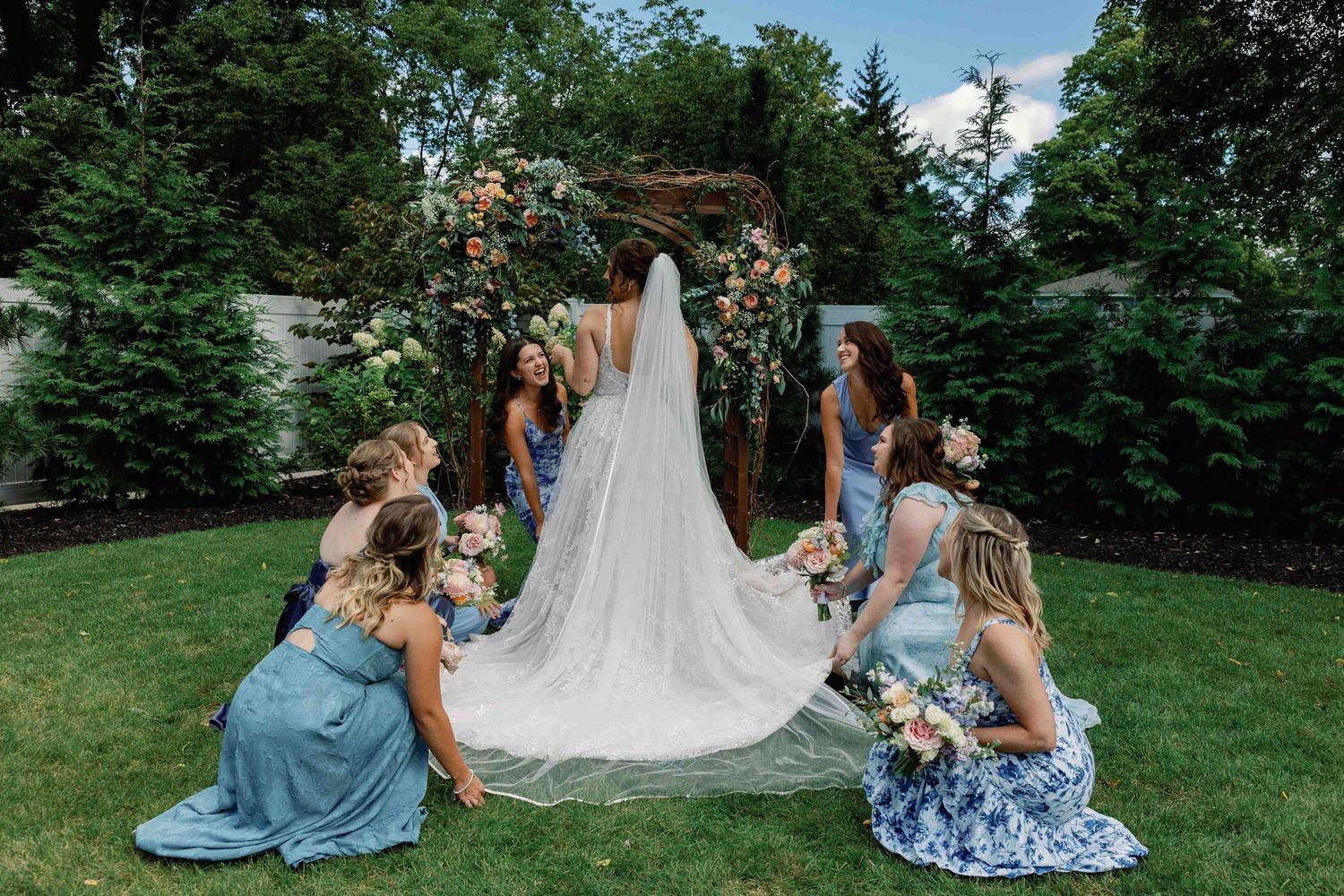 Bridesmaids in blue dresses arrange a long white wedding veil in an outdoor garden setting.