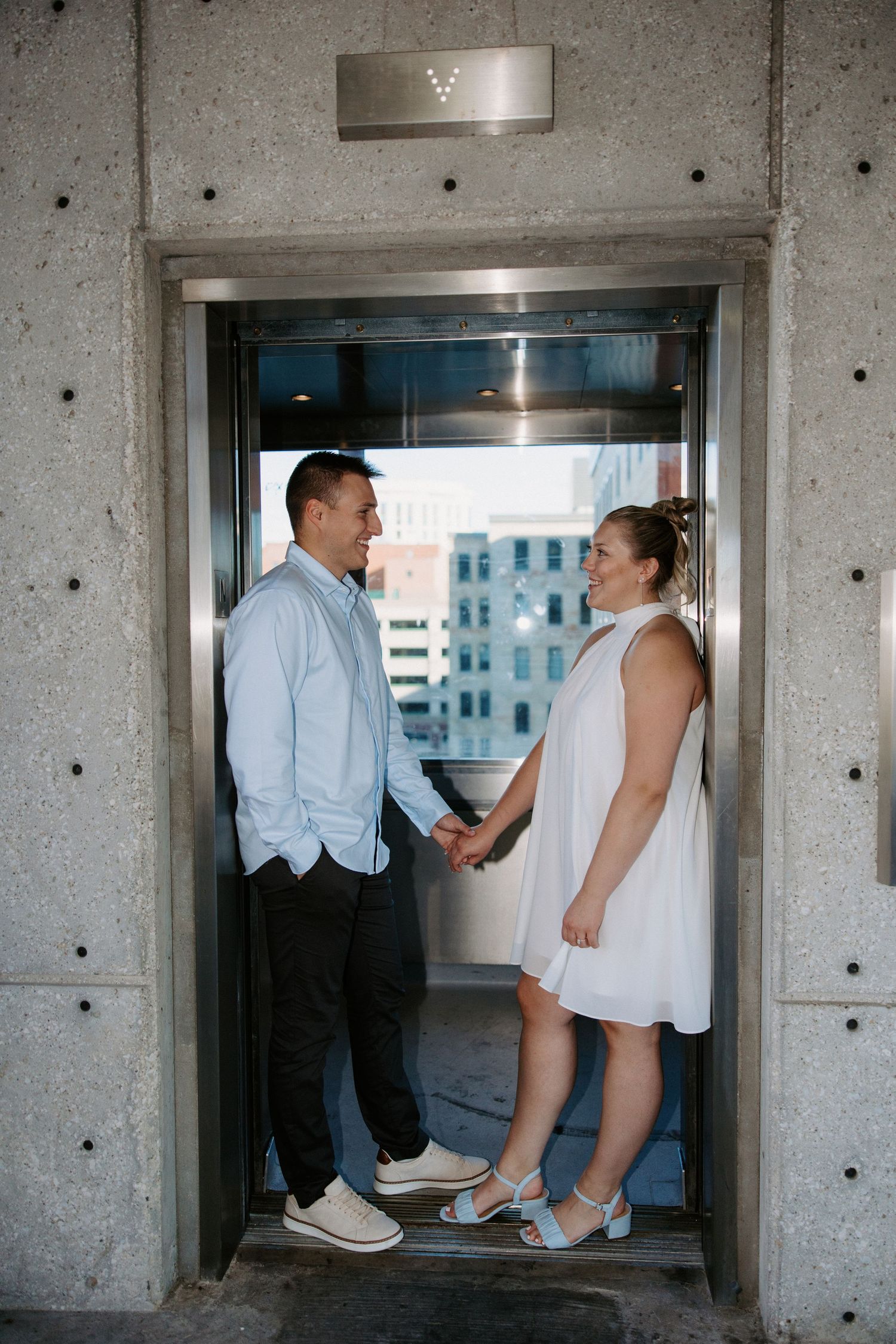 A couple in light summer attire poses together in an industrial elevator setting.