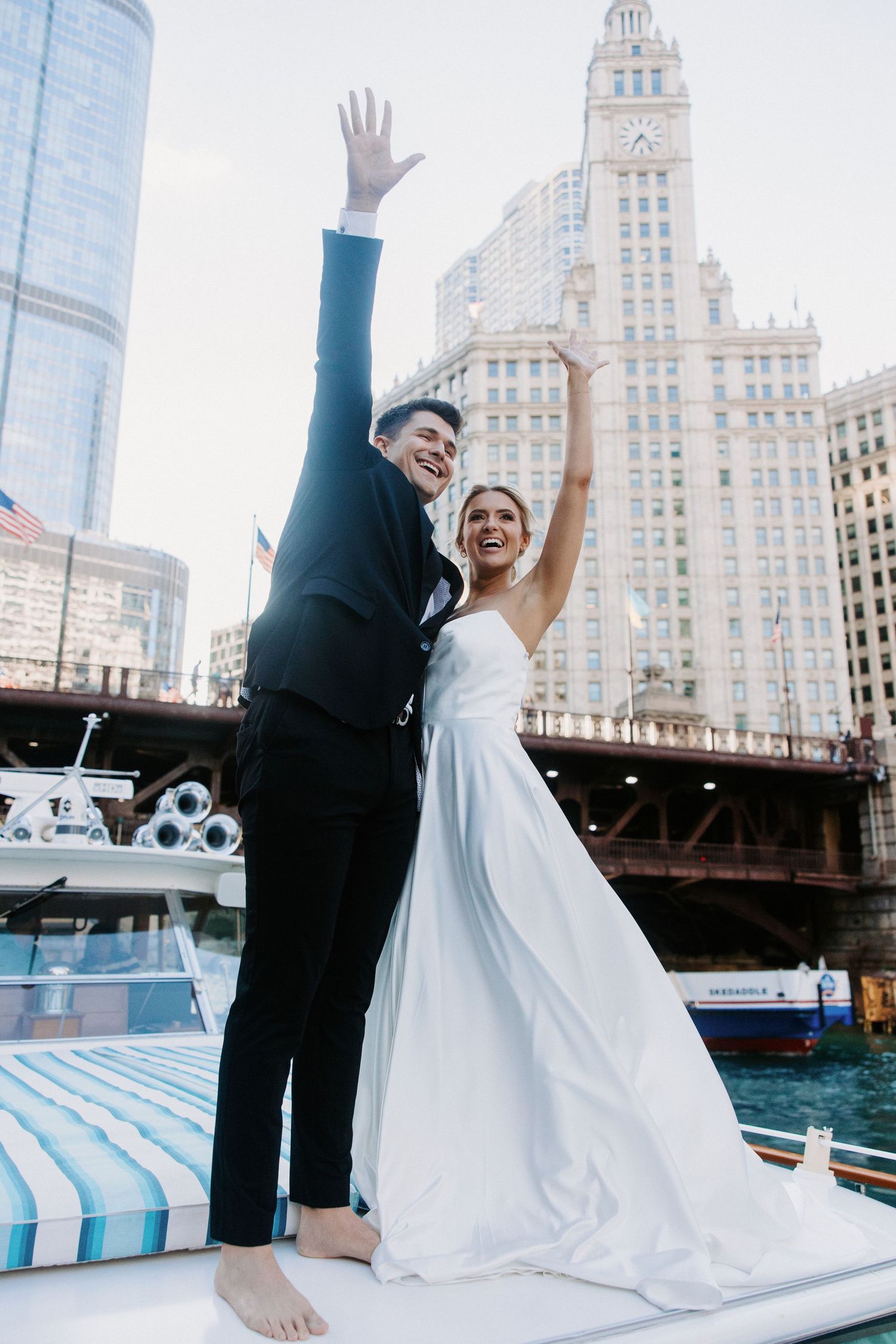 Joyful wedding couple celebrating with raised arms on Chicago riverboat with Wrigley Building visible.