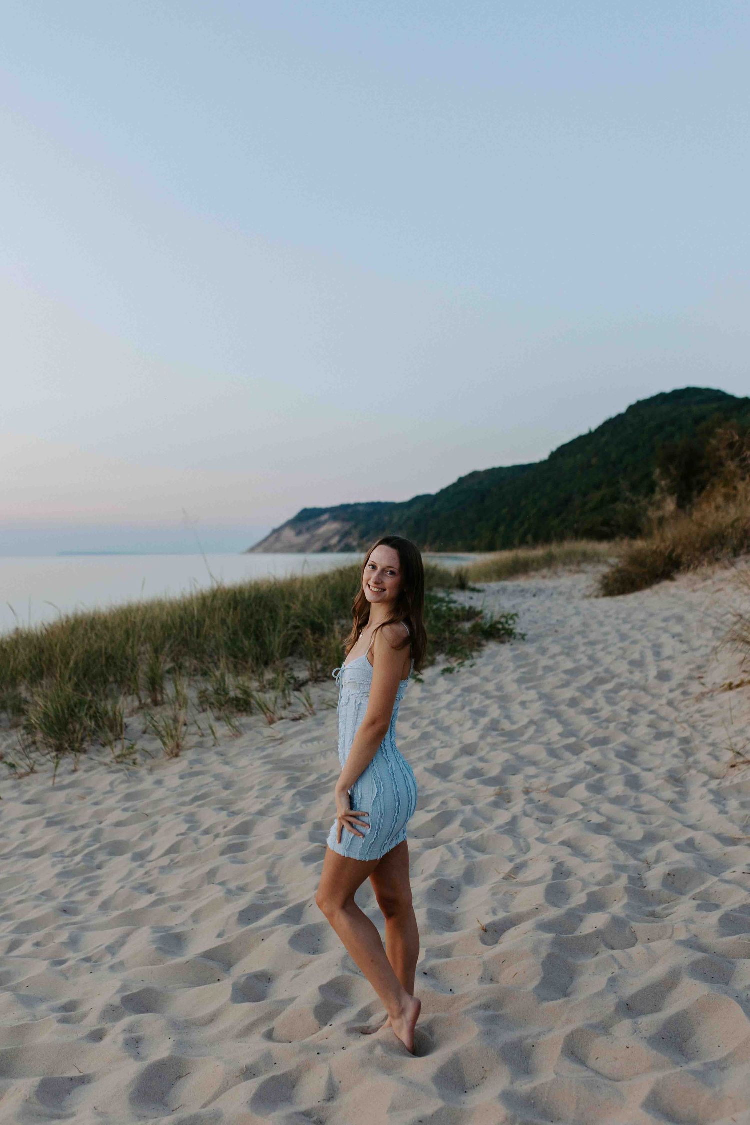 Figure in a blue dress poses gracefully on sandy beach dunes during golden hour.
