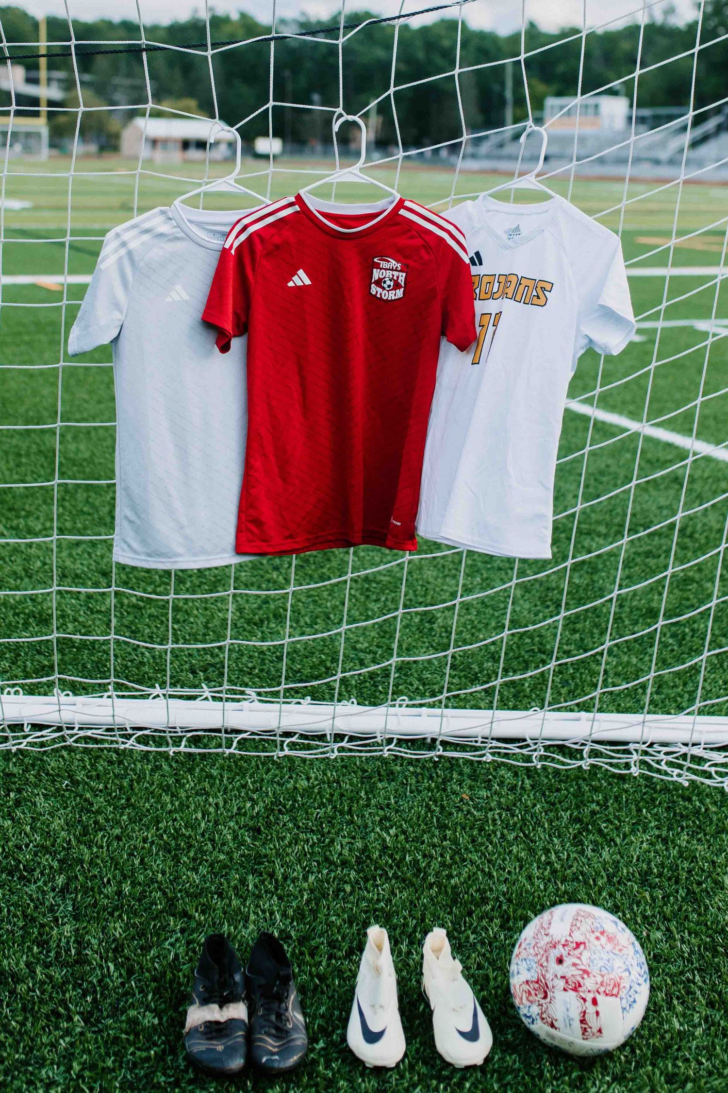 Soccer jerseys, cleats and balls displayed along goal net on artificial turf field.