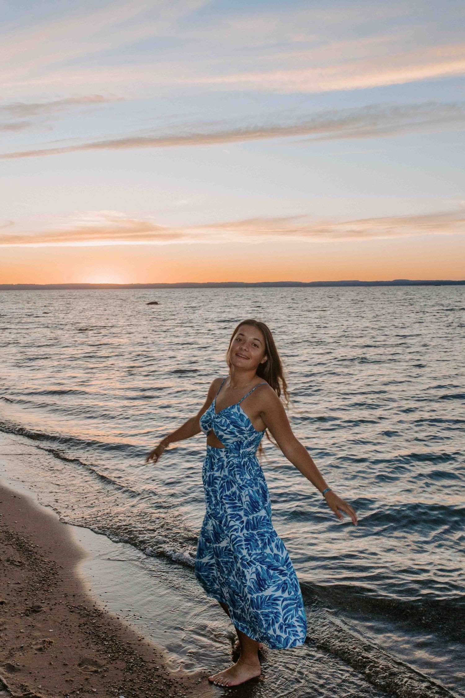 Person in blue dress dances playfully on beach shoreline during sunset.