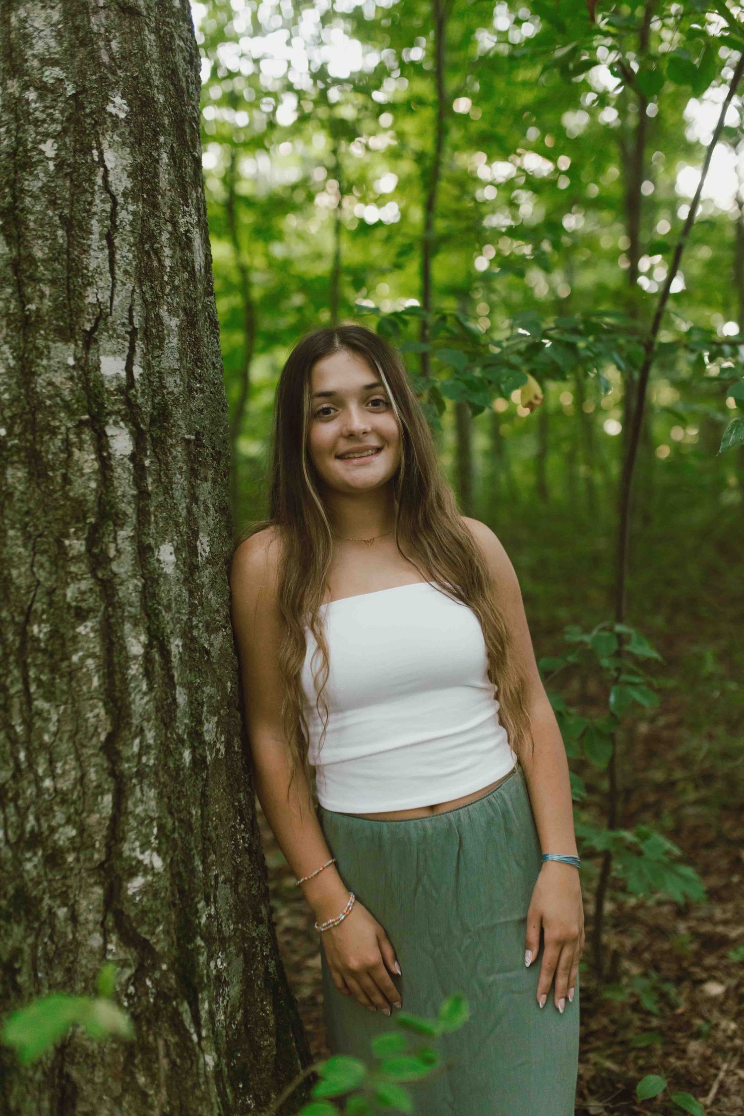 A summer photoshoot in a wooded setting with white top and olive skirt against tree trunks.