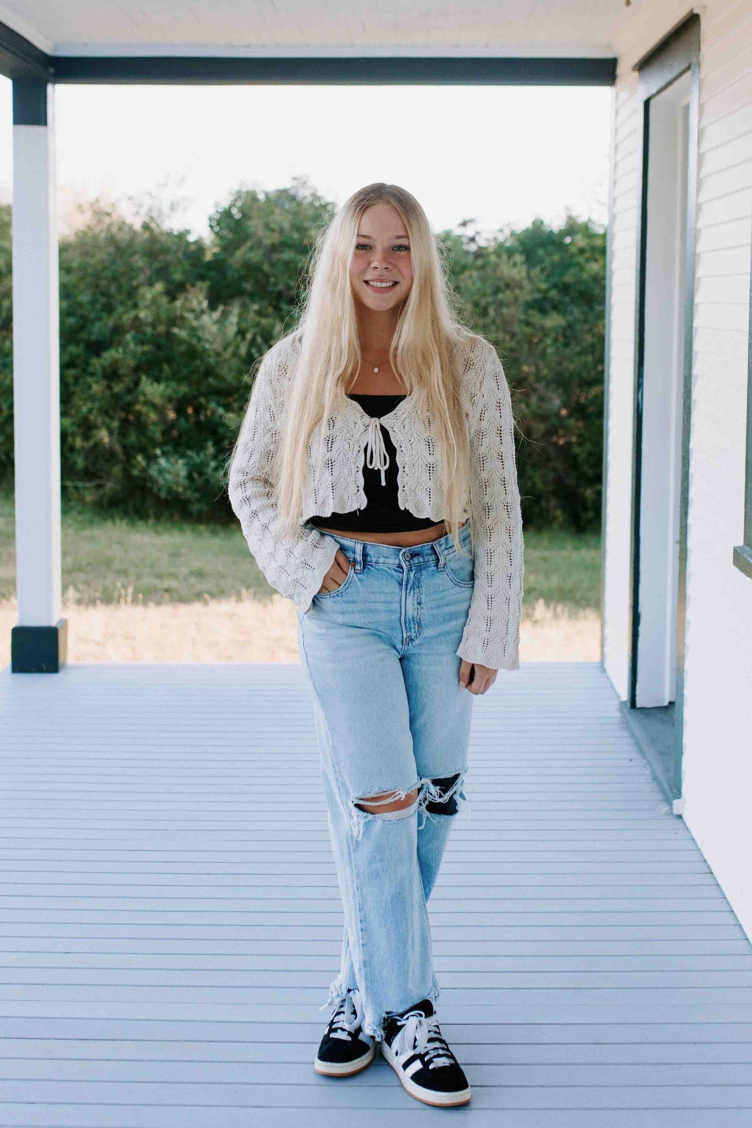 A relaxed pose on a white porch wearing distressed jeans, a crochet sweater and casual footwear.