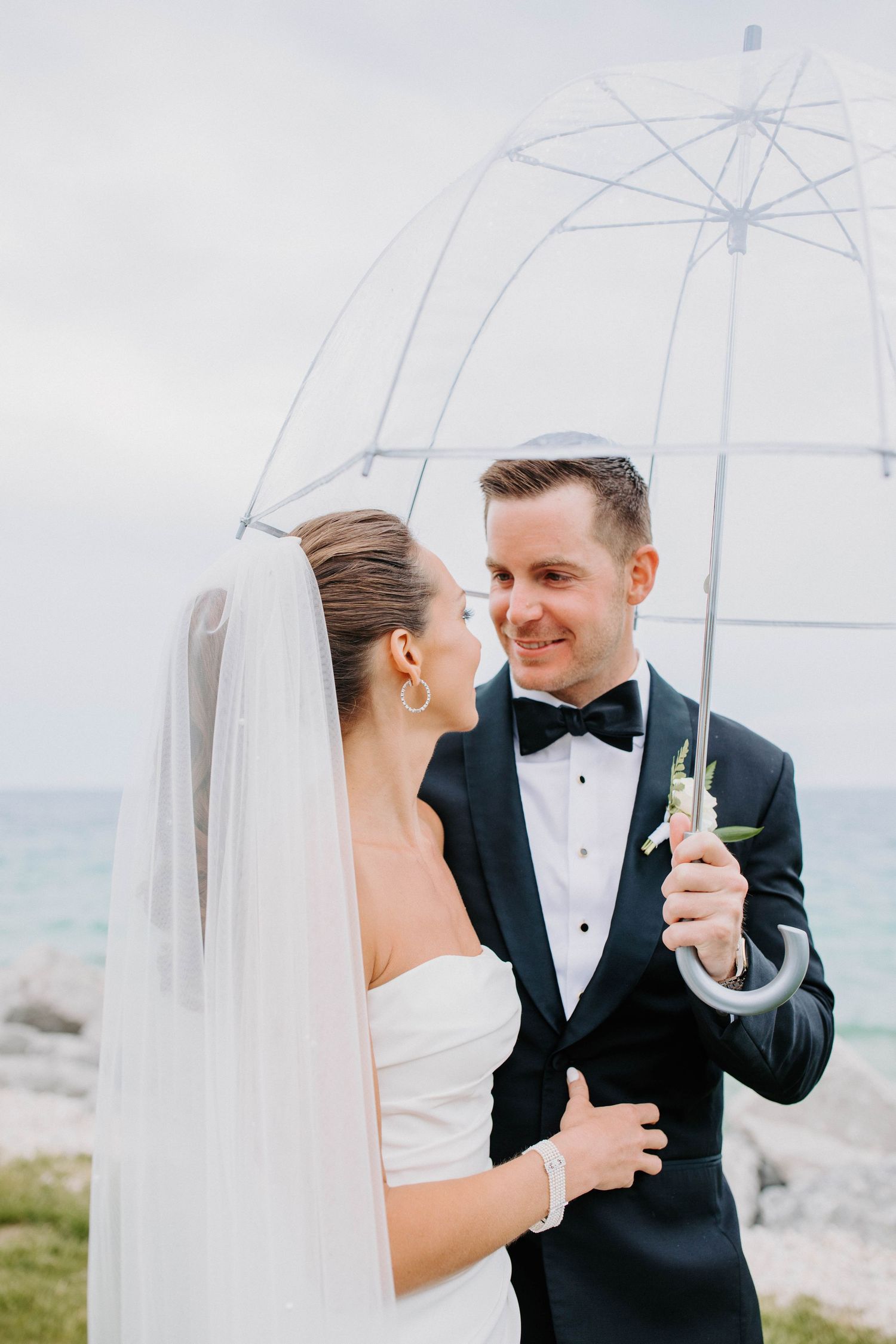 A wedding couple shares a romantic moment under a clear umbrella by the ocean.