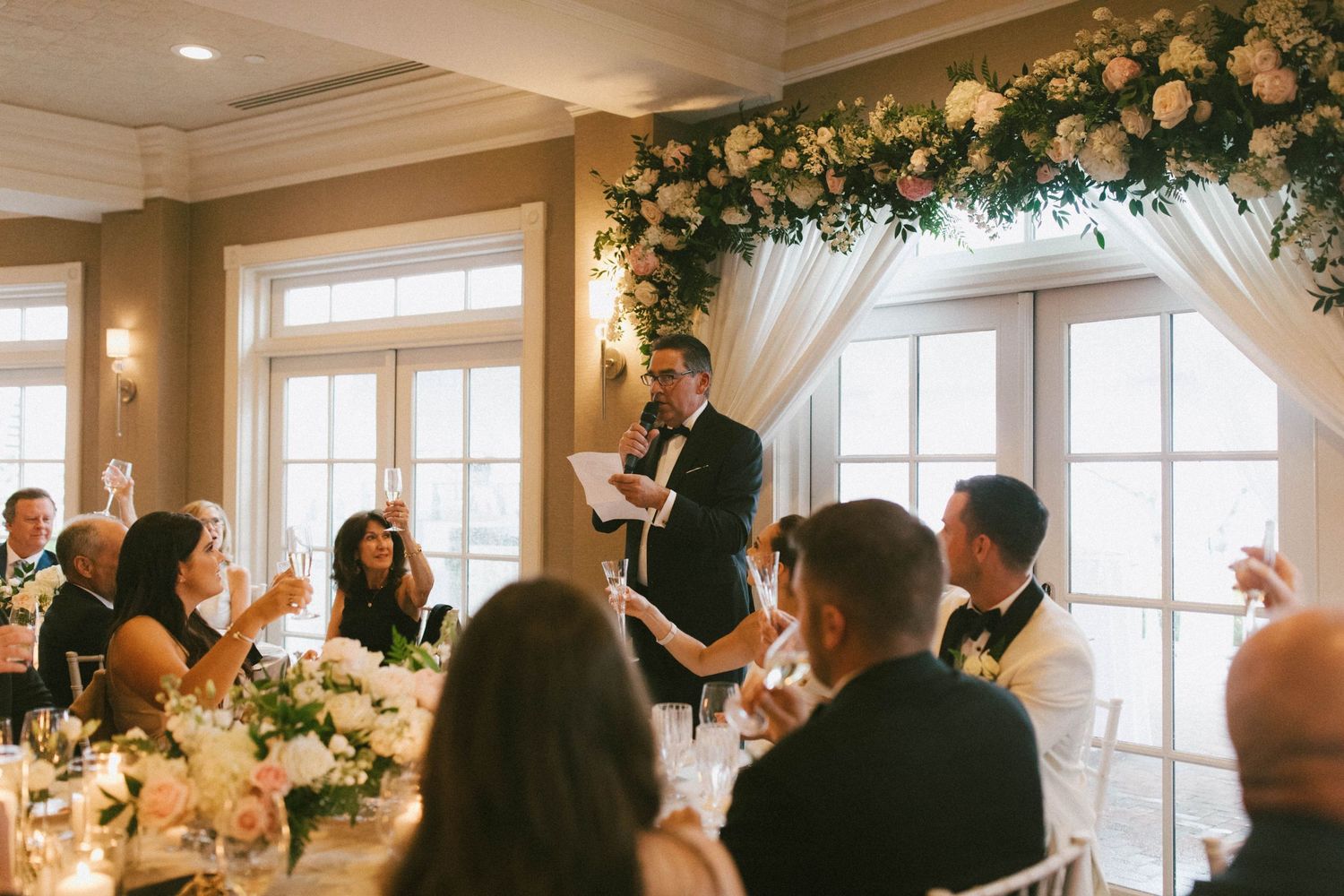Wedding toast being given under an elegant floral arch with white draping and natural light.