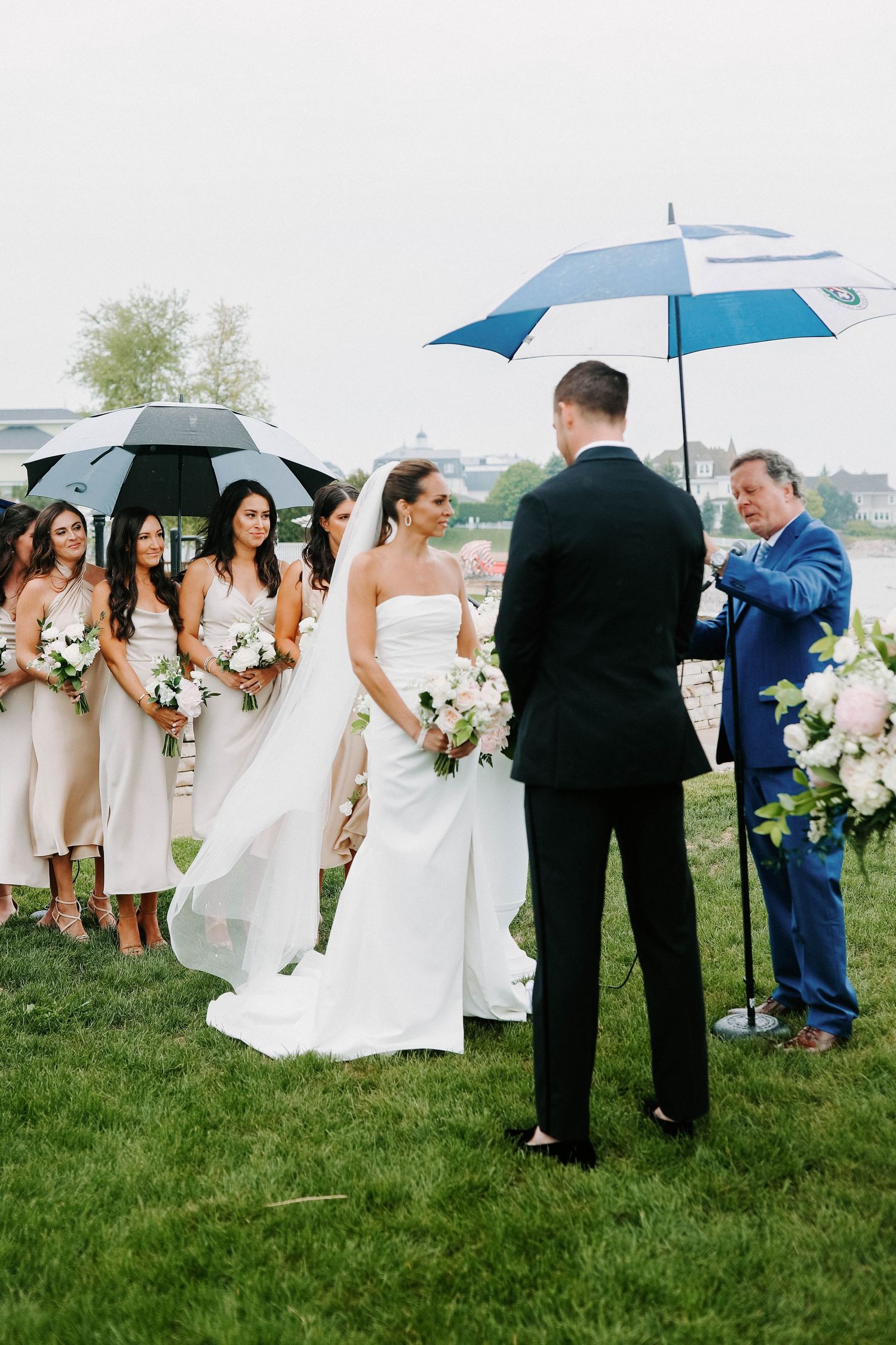 Wedding ceremony participants stand on grass holding umbrellas near elegant white floral arrangements.