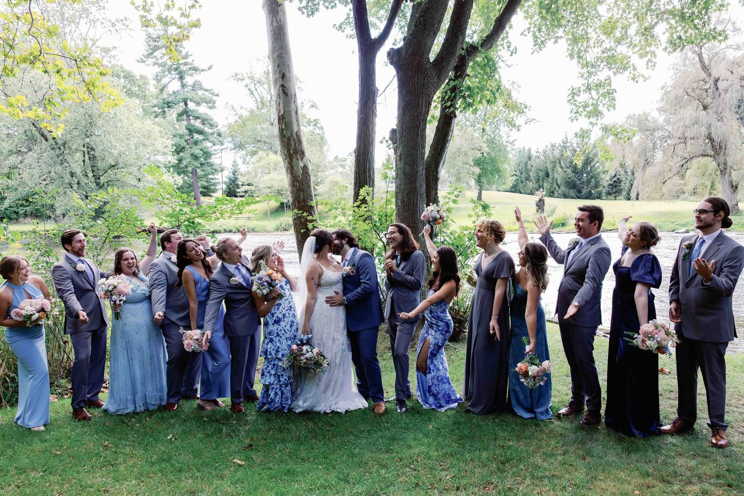 Large wedding party lines up for a group photo in a natural outdoor setting with trees in the background.