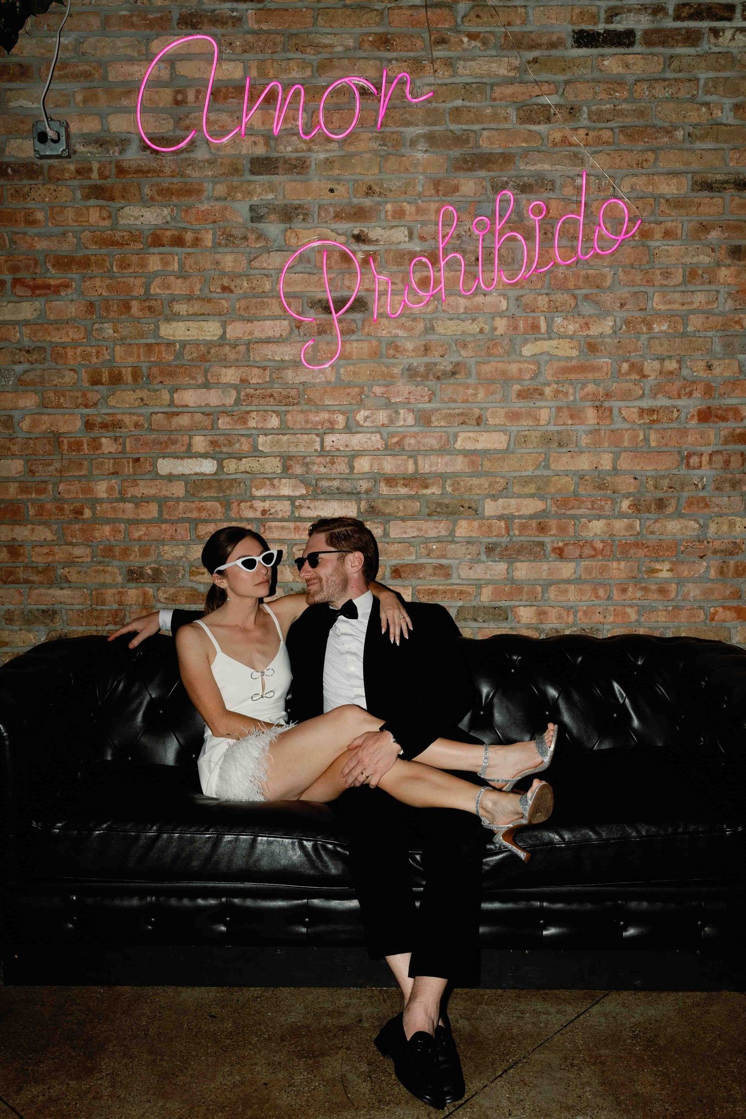 Couple relaxes on black leather couch under pink neon signs at venue.