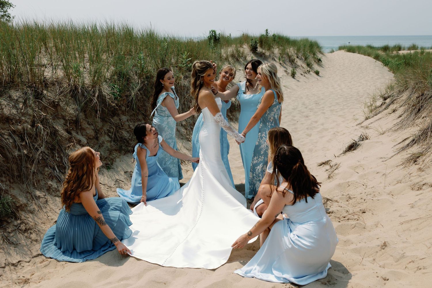 Wedding party members in coordinated blue dresses pose together on sandy beach dunes.