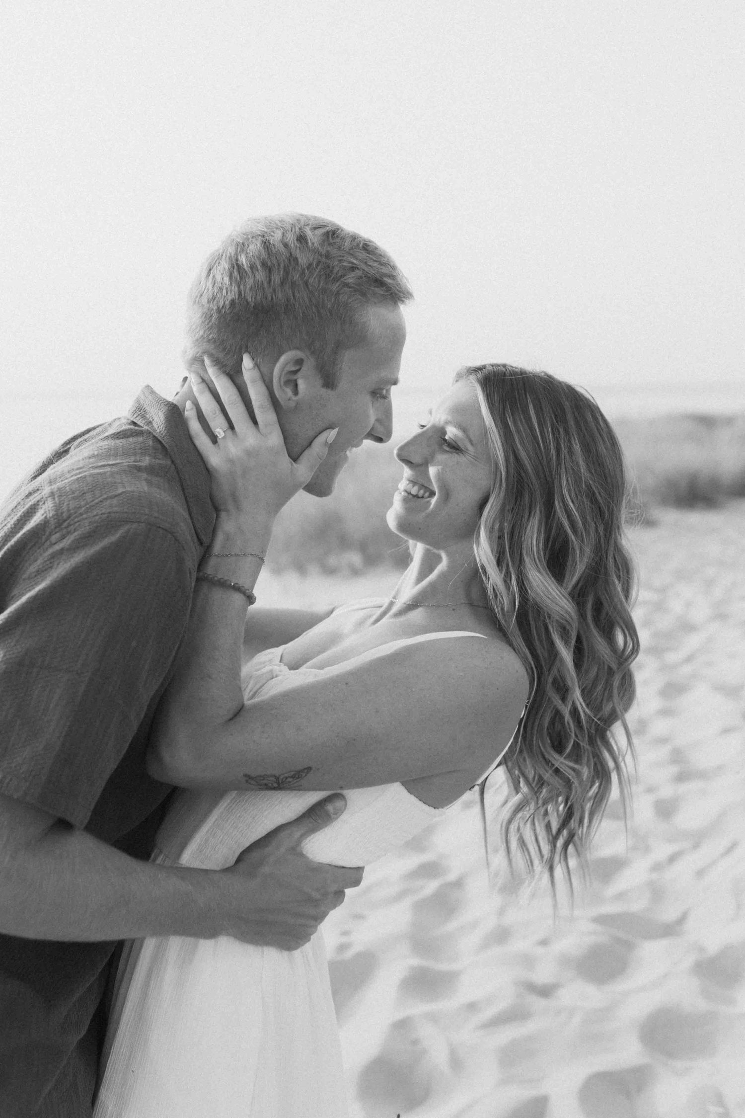A romantic black and white beach photo of a couple embracing in the sand with soft lighting.