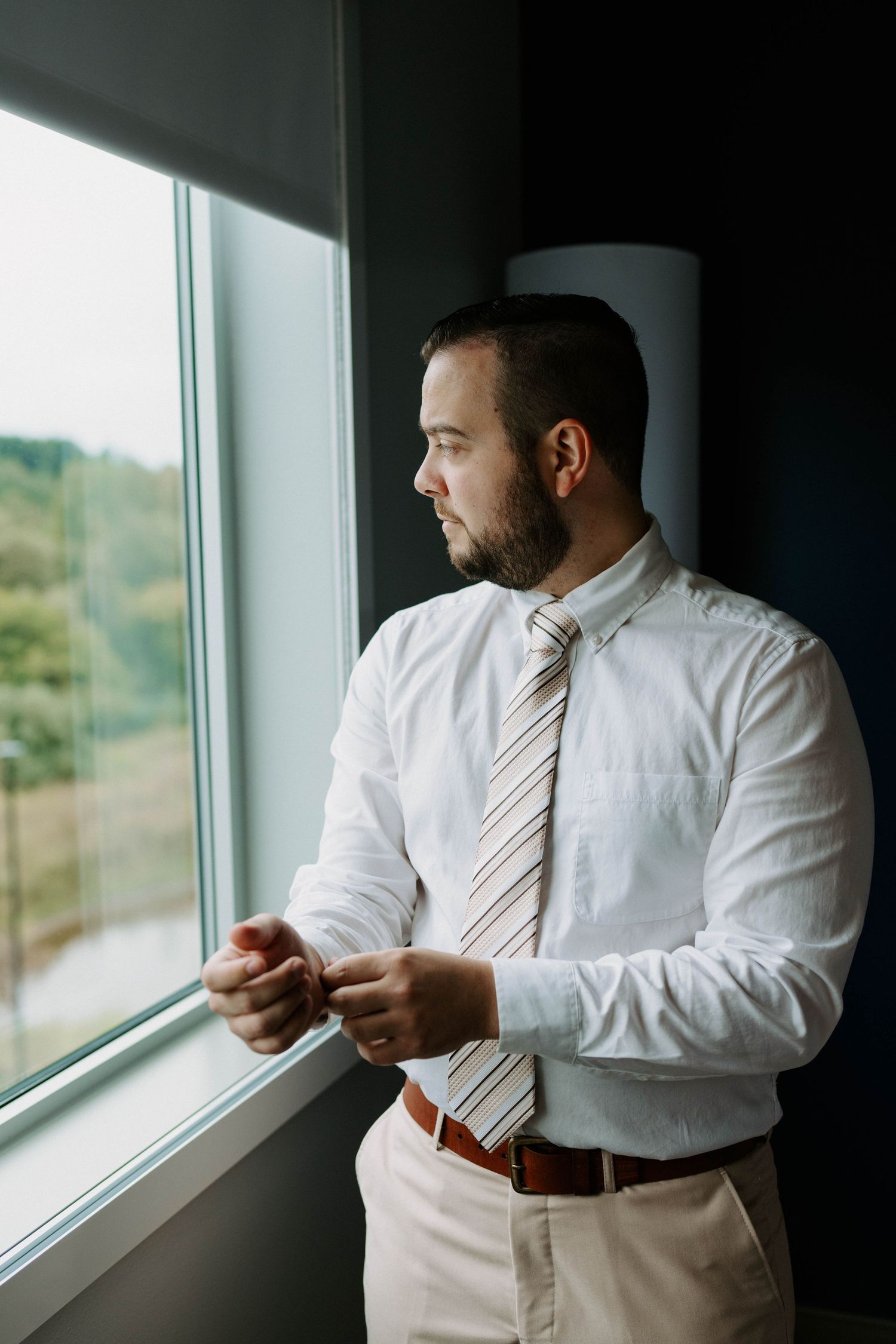 Groom in formal attire adjusts his tie while looking out large windows.