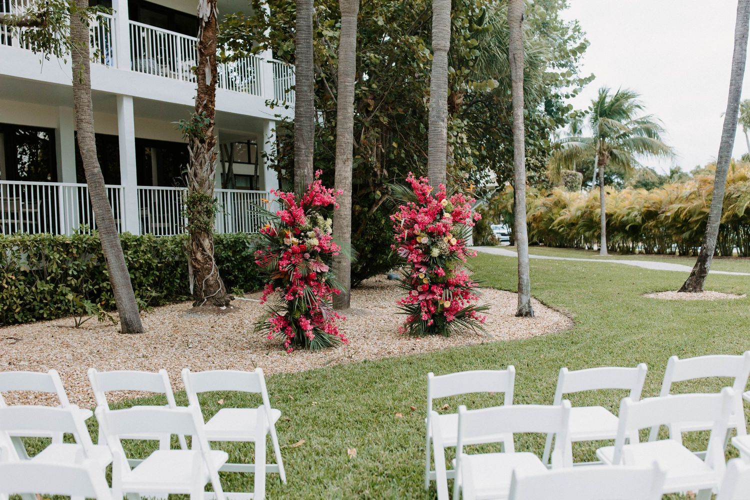 White wedding chairs arranged in rows on a manicured lawn with palm trees and pink bougainvillea flowers.