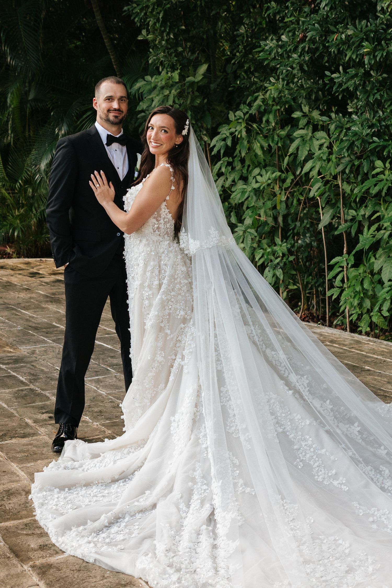 Bride in stunning lace gown with cathedral train poses on stone patio surrounded by lush greenery.