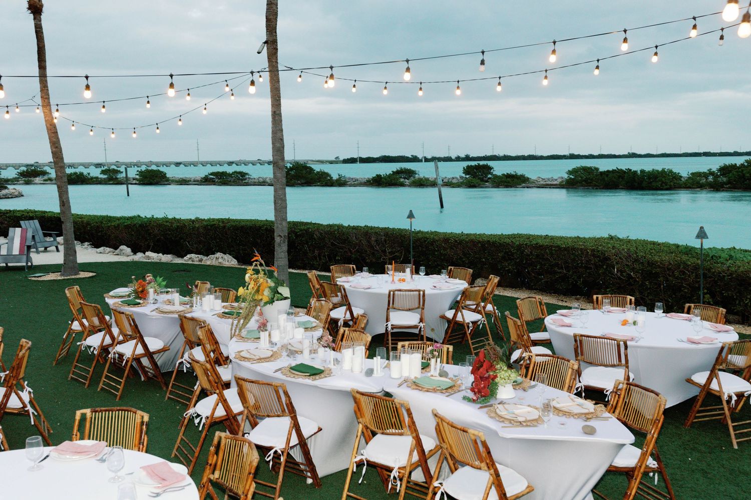 An outdoor wedding reception setup with string lights and round tables overlooking the water.