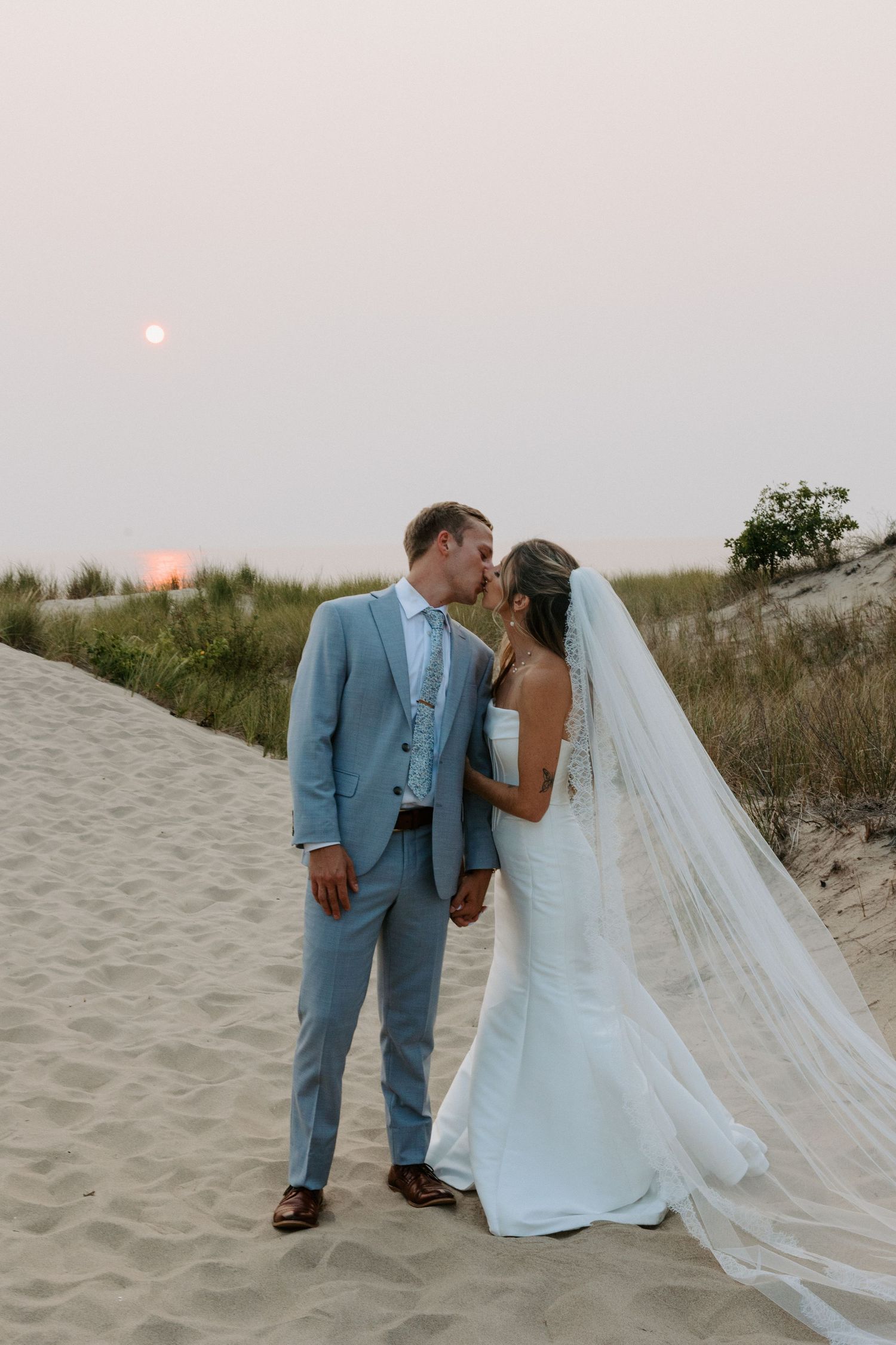 Sunset beach wedding portraits with soft lighting and flowing white veil against sand dunes.