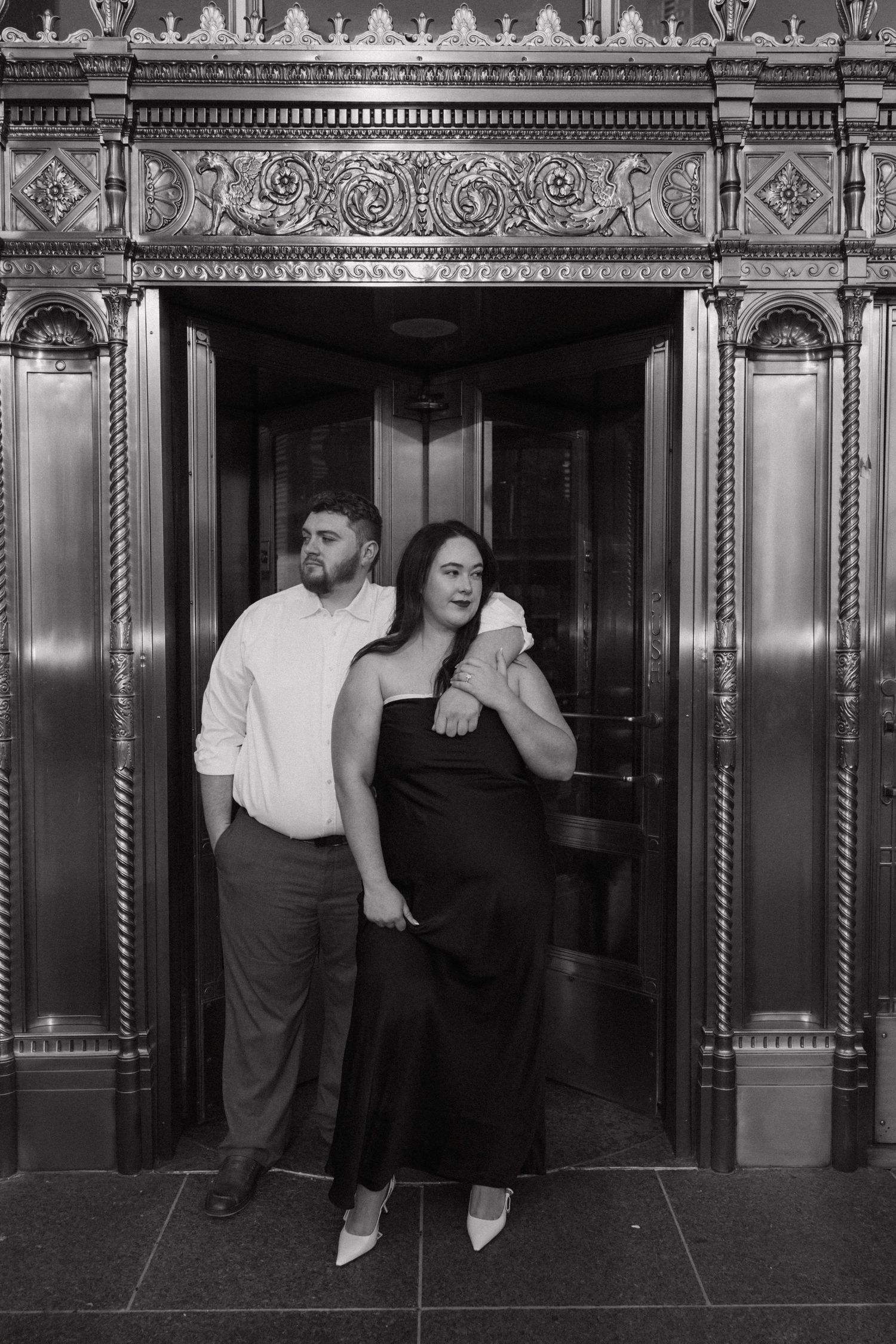 A couple poses against vintage elevator doors with ornate metalwork in black and white.