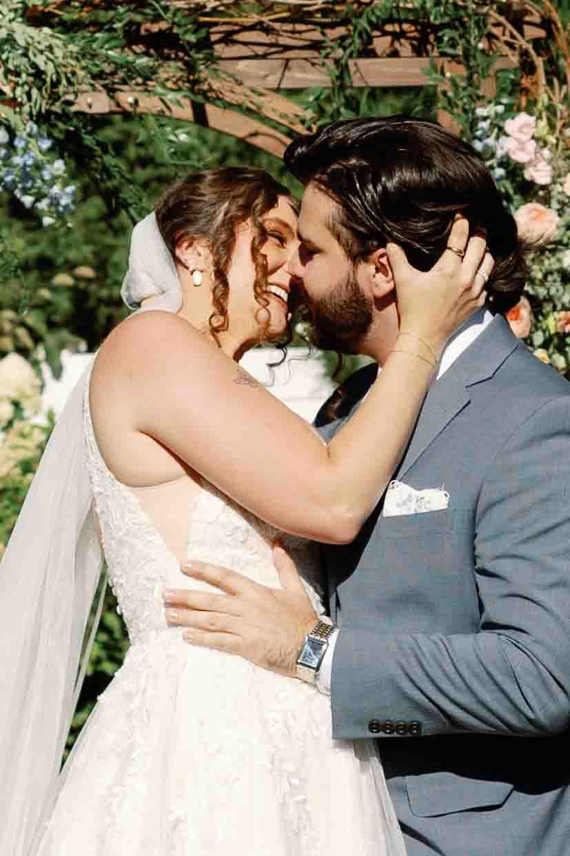 Newlyweds share their first kiss as a married couple under a floral arch outdoors.