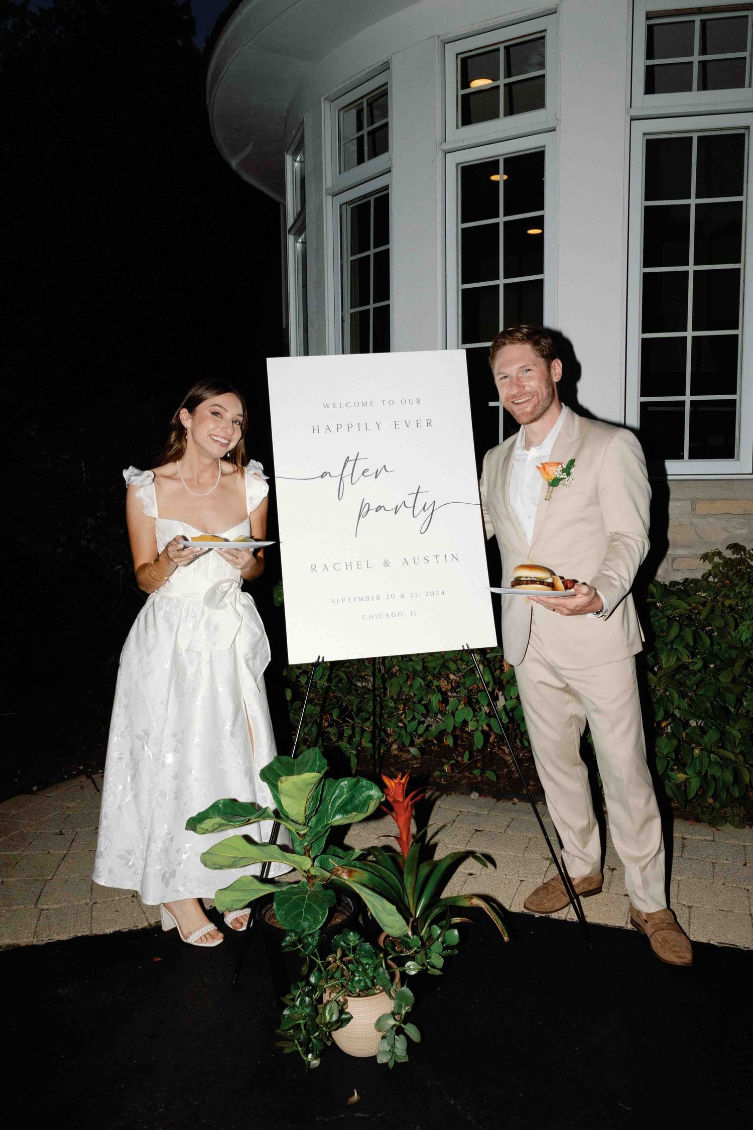 Wedding couple poses with signs outside white building at night.