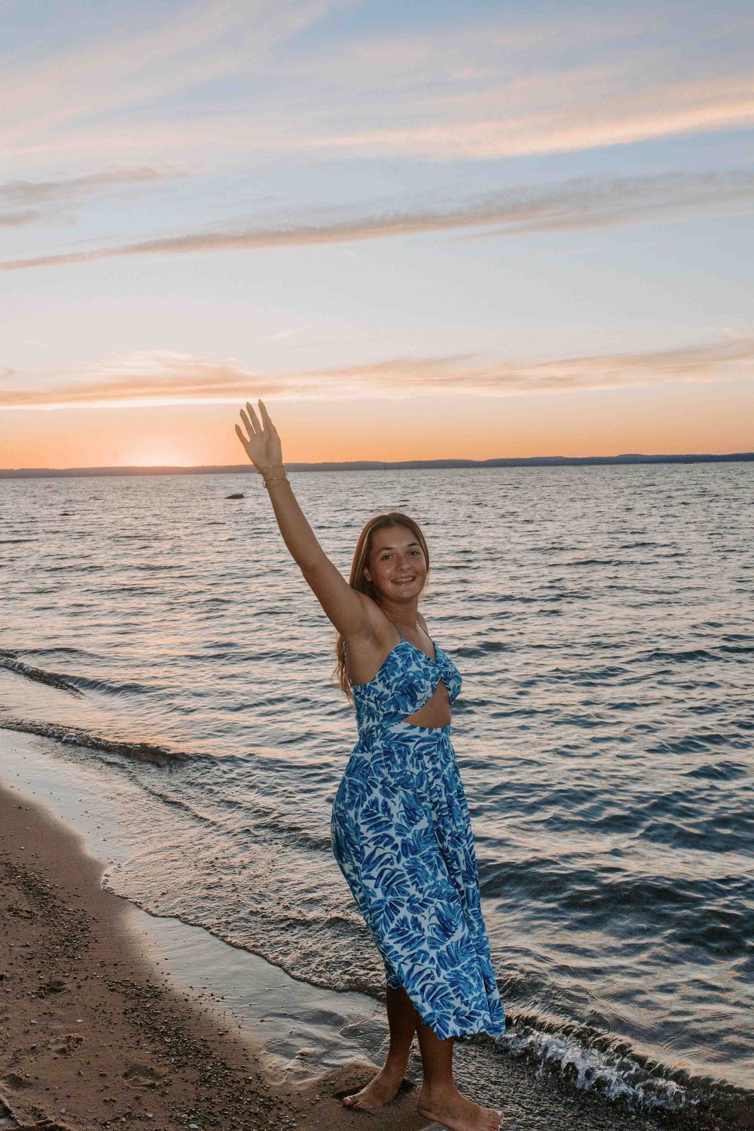Person in blue dress waves happily on beach with orange sunset sky.