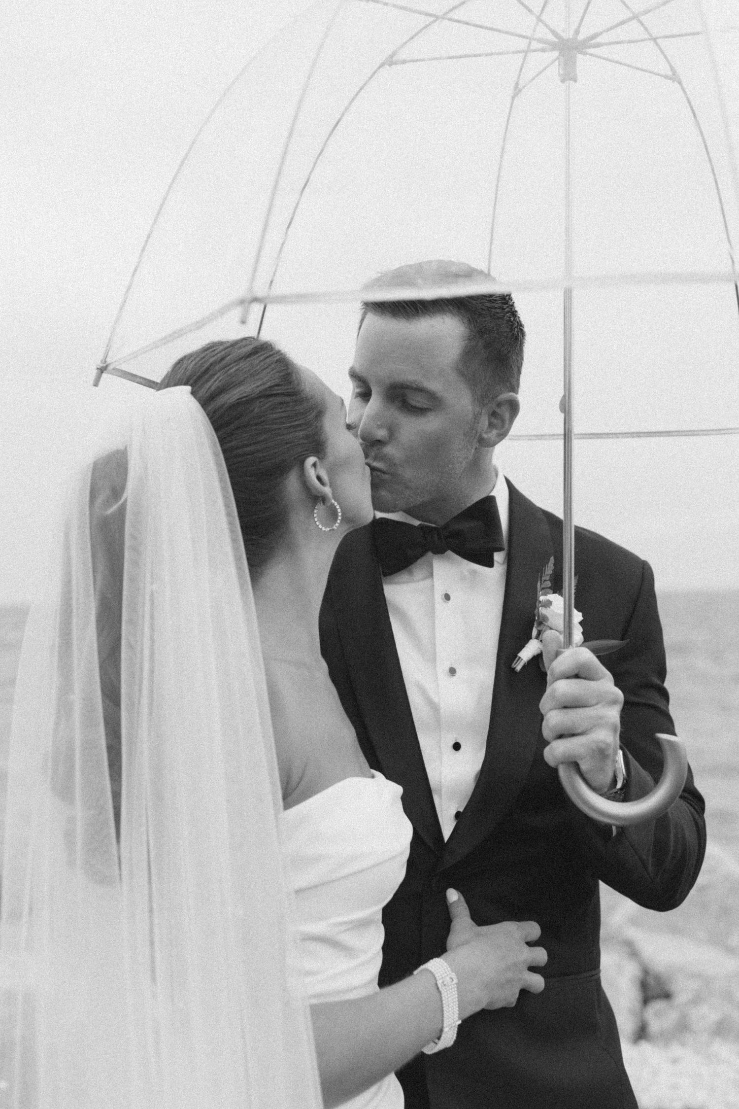A black and white portrait of newlyweds sharing an intimate moment under a clear umbrella.