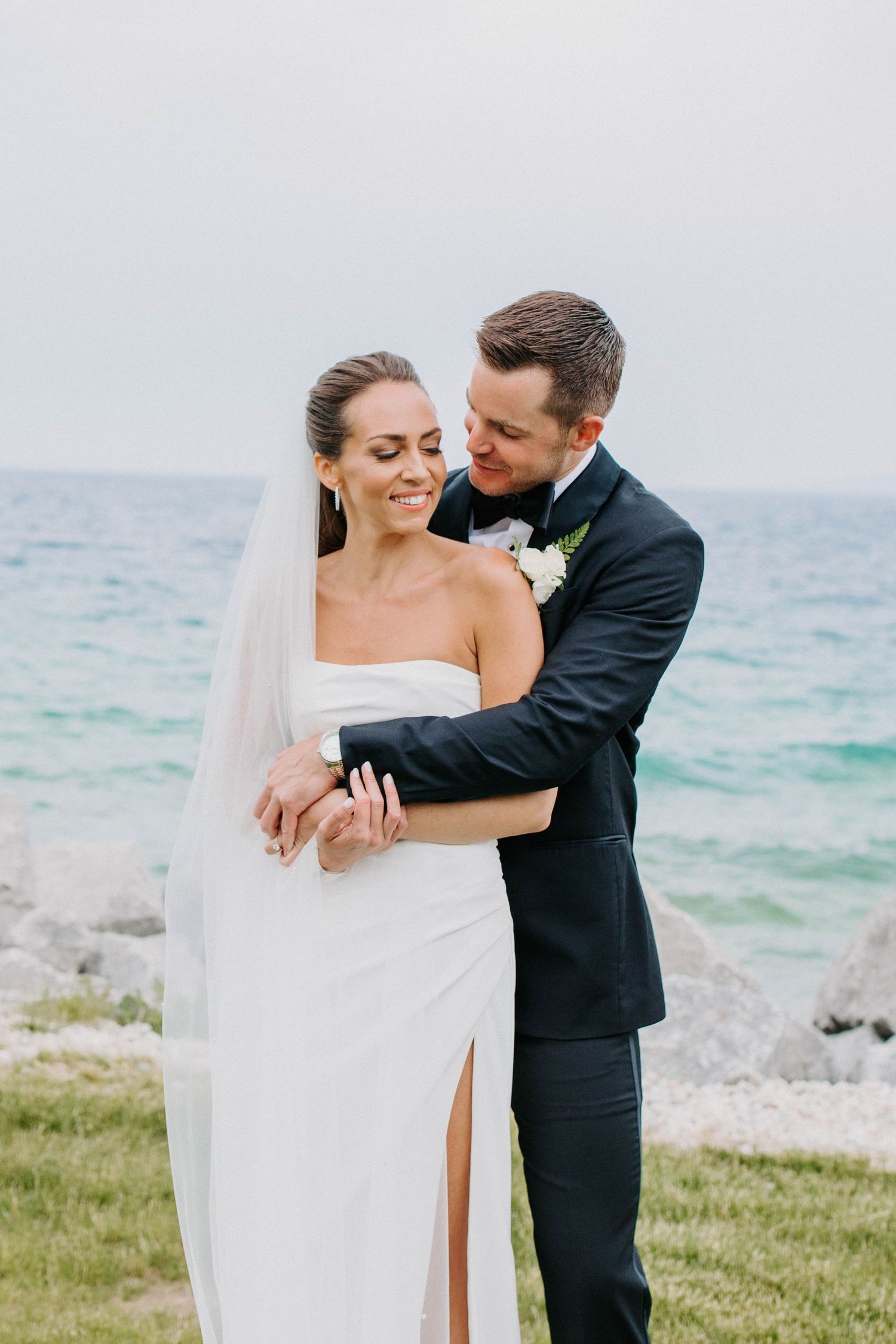 Romantic wedding portrait by the ocean showing couple embracing on grass near rocky shore.