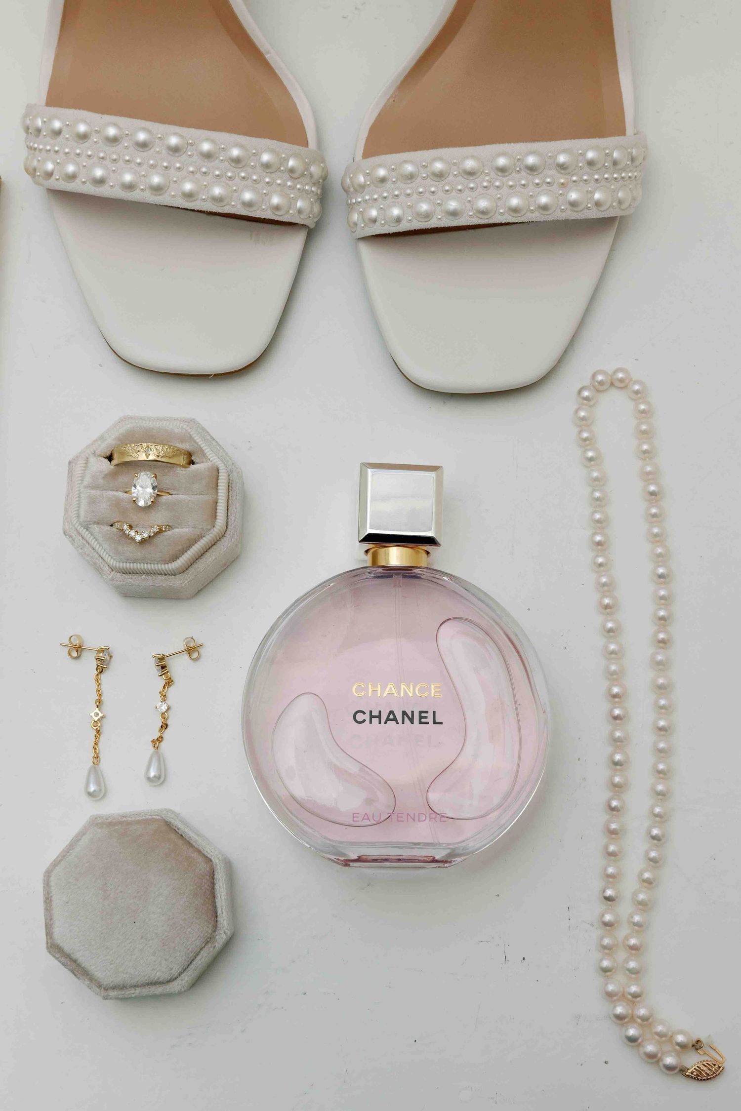 Wedding accessories including pearl jewelry, white sandals, and pink Chanel perfume bottle arranged on white surface.