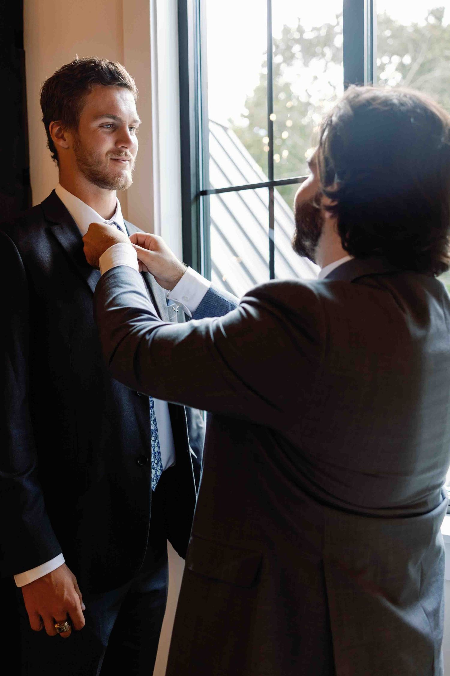 Person adjusting another's tie by window in formal wedding attire.
