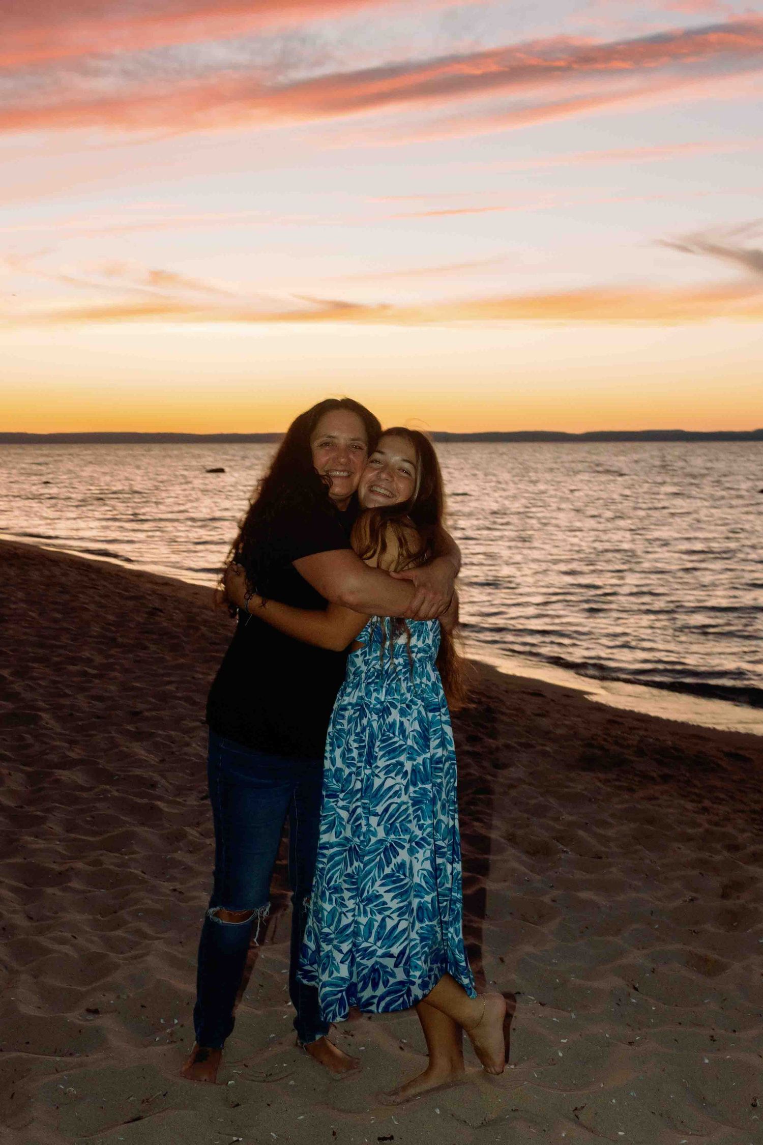 People embracing on sandy beach at sunset with colorful sky and ocean backdrop.