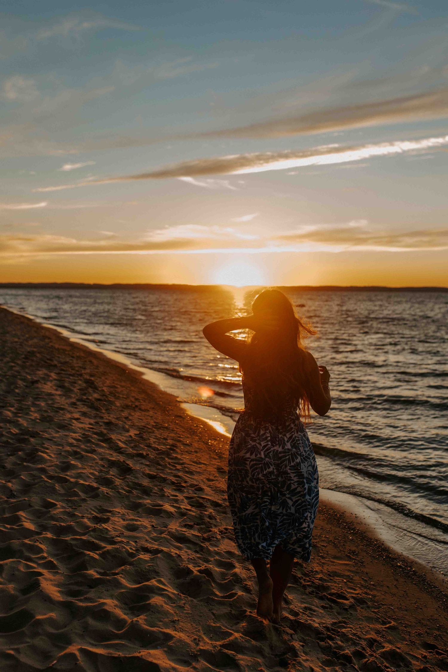Silhouette figure in blue dress stands on sandy beach during golden sunset over calm ocean waters.