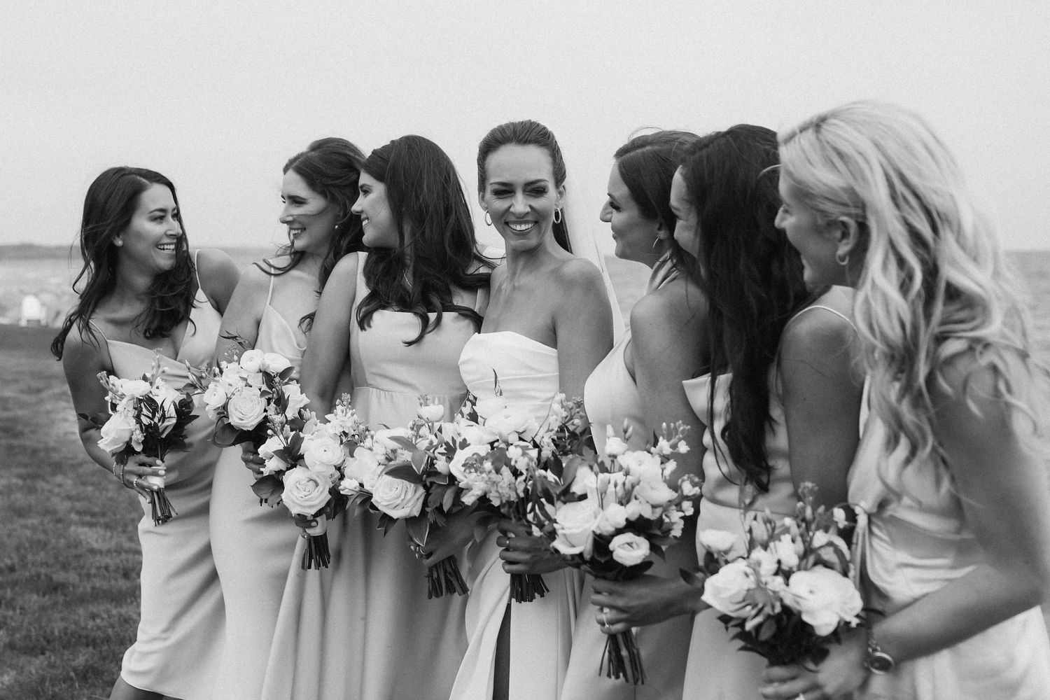 Black and white photo of wedding party holding bouquets and sharing candid moment.