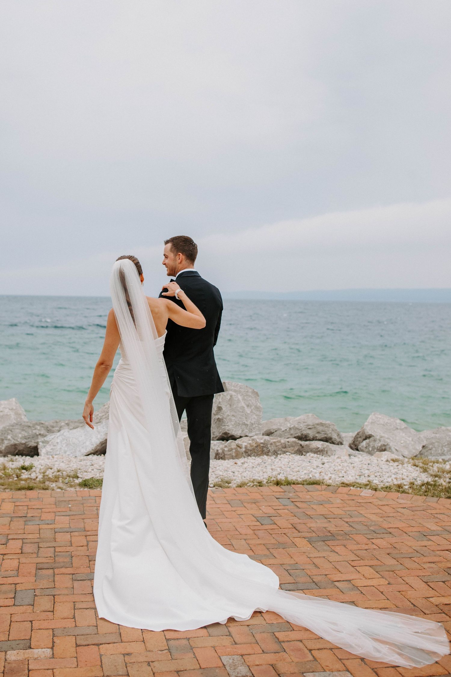 A bride's flowing white dress train spreads across red brick pavers by the ocean.