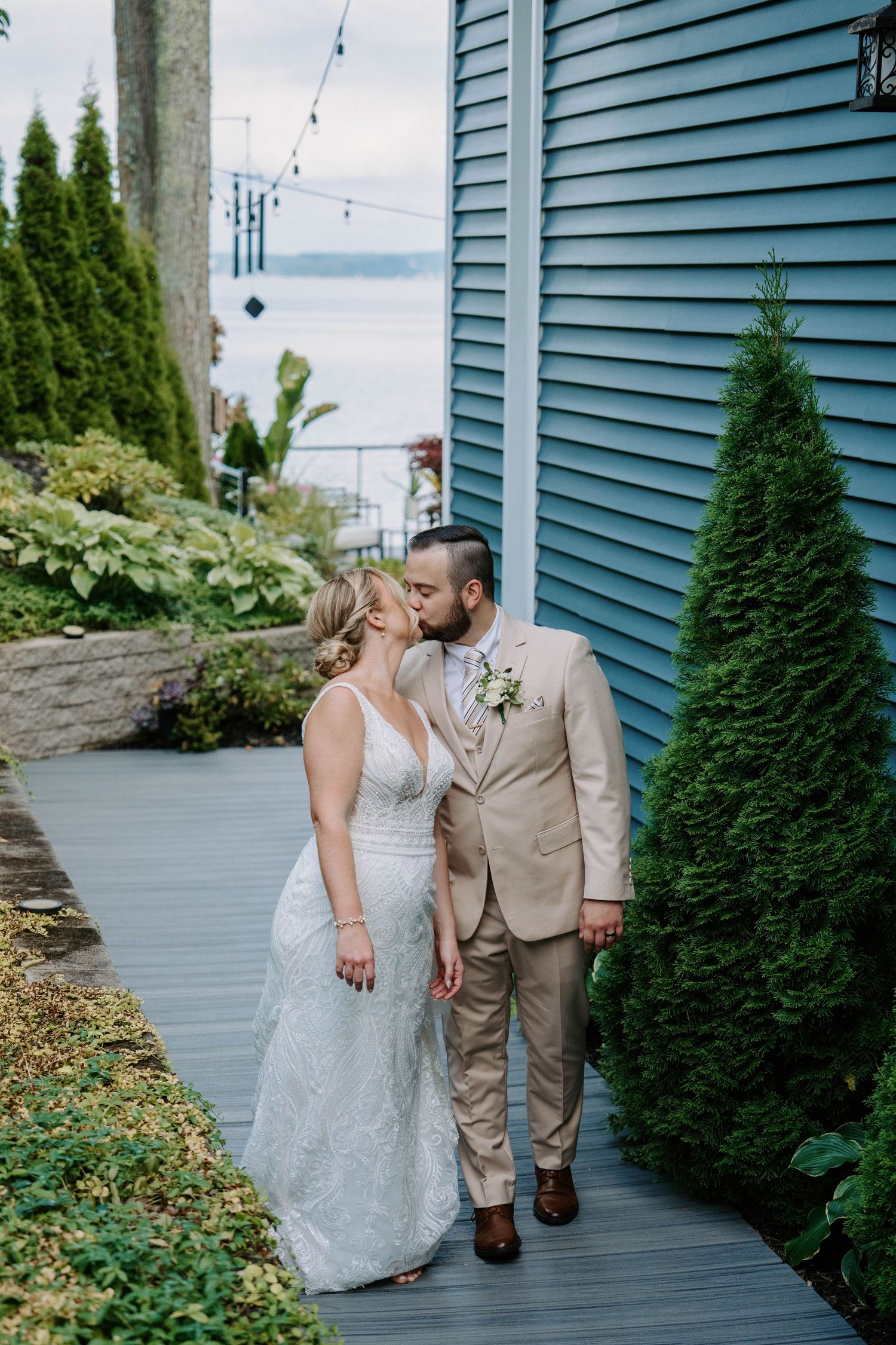 A couple in wedding attire shares a romantic moment on a wooden deck surrounded by evergreen shrubs.