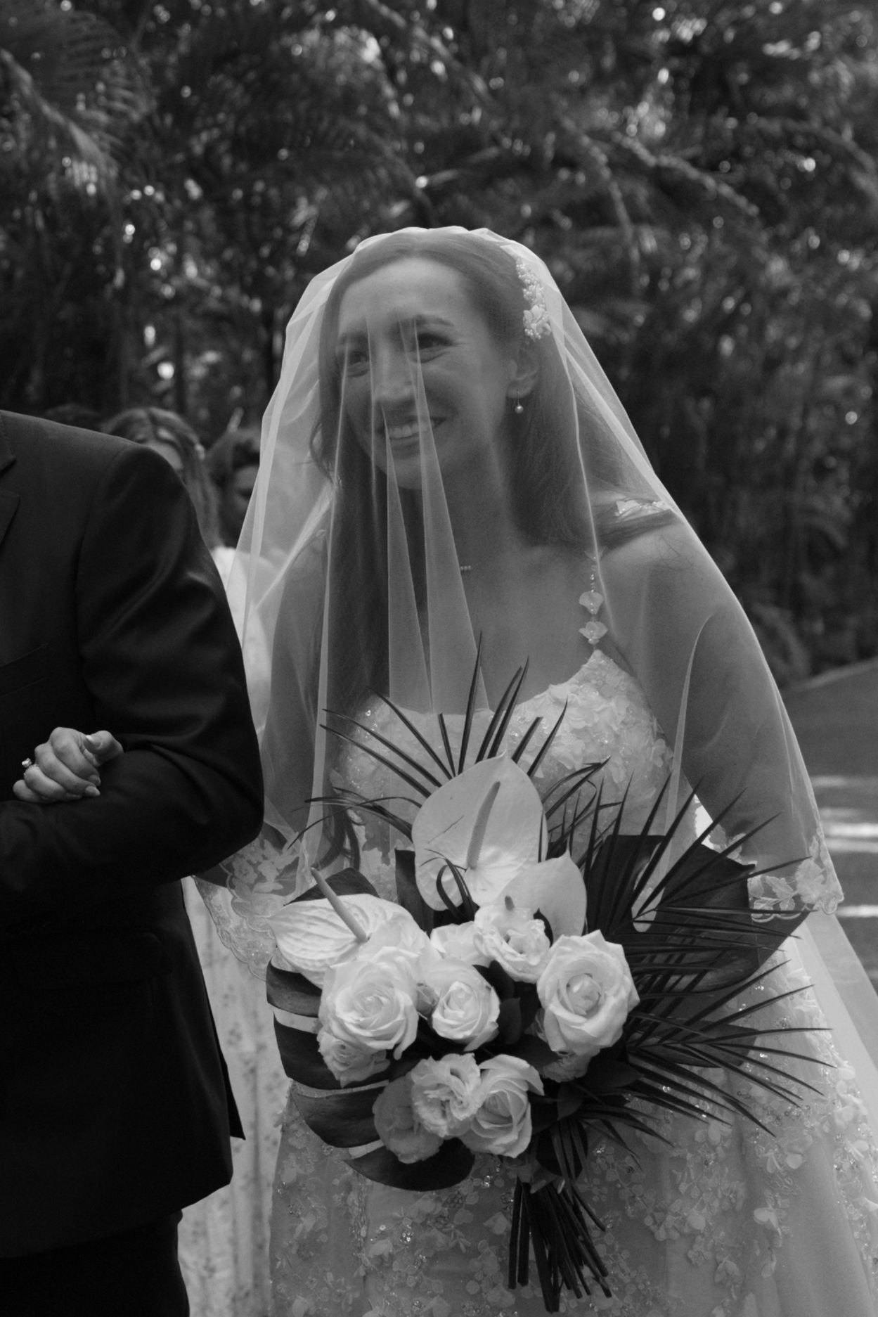 Bridal bouquet features white roses and tropical palm leaves against lace wedding dress.