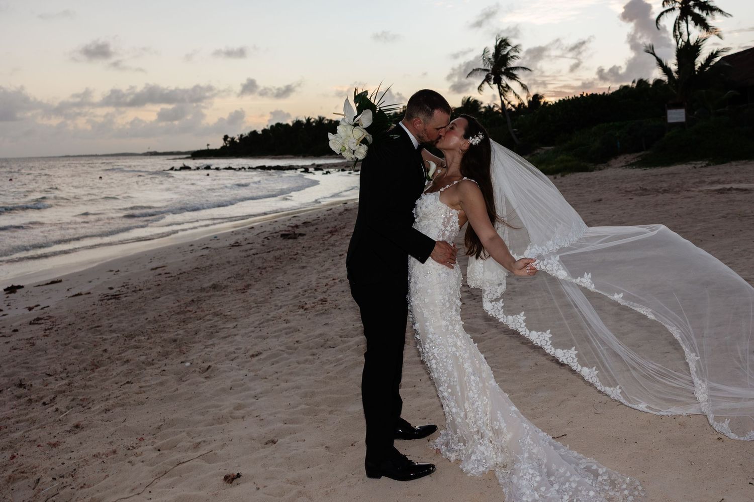 A romantic beach wedding moment at sunset with palm trees and flowing veil.