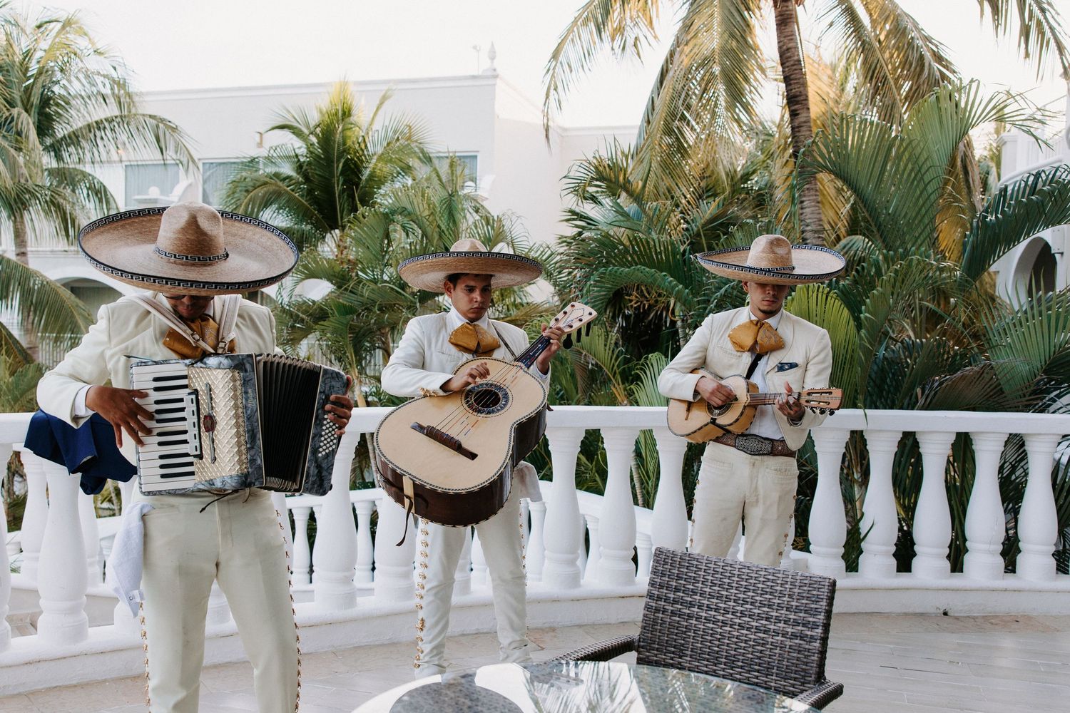 Three mariachi musicians perform on a white balcony with palm trees in background.