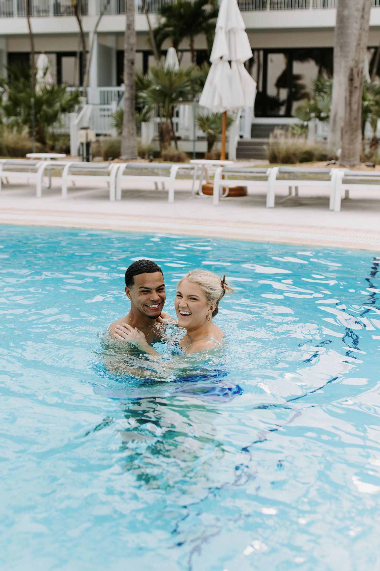 Joyful couple enjoying a swim in a resort pool with lounge chairs in background.
