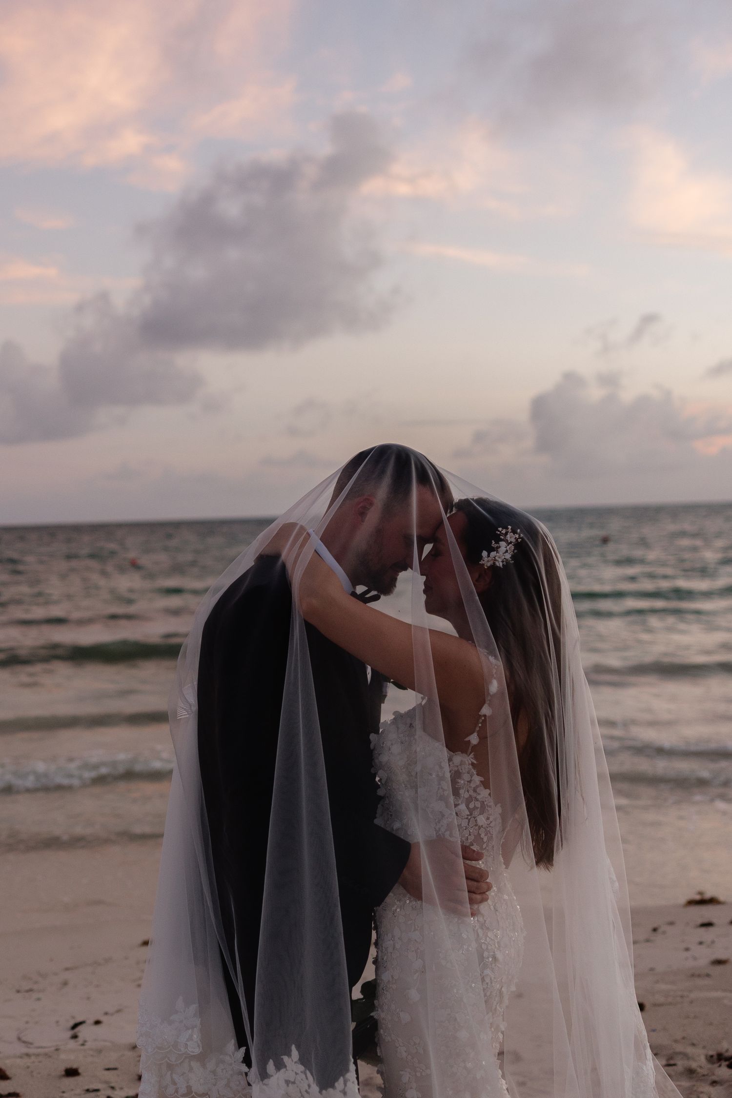 A romantic sunset beach moment with flowing veil and soft pink clouds.