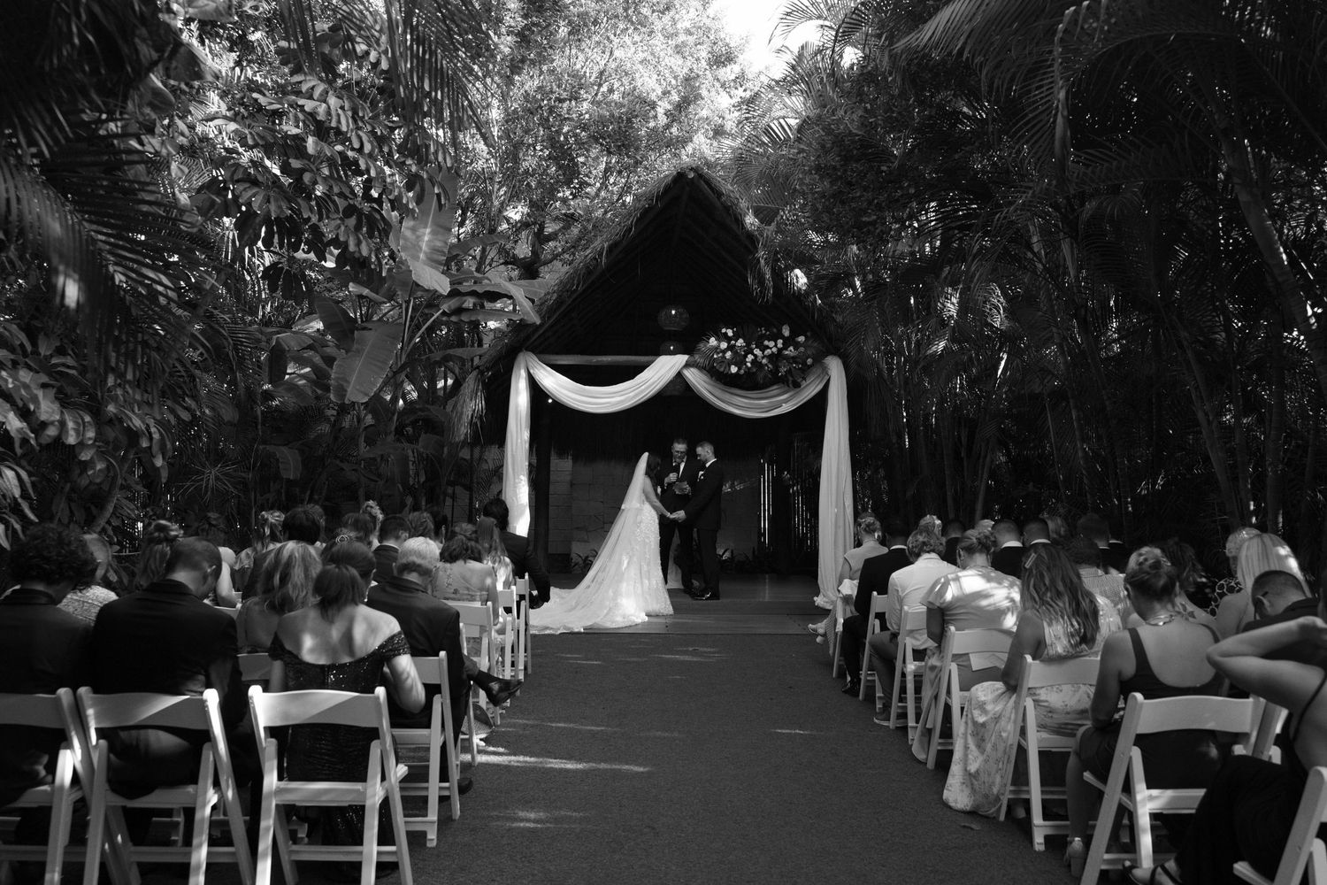 Wedding ceremony setup with white chairs and floral decorations along tree-lined outdoor path.