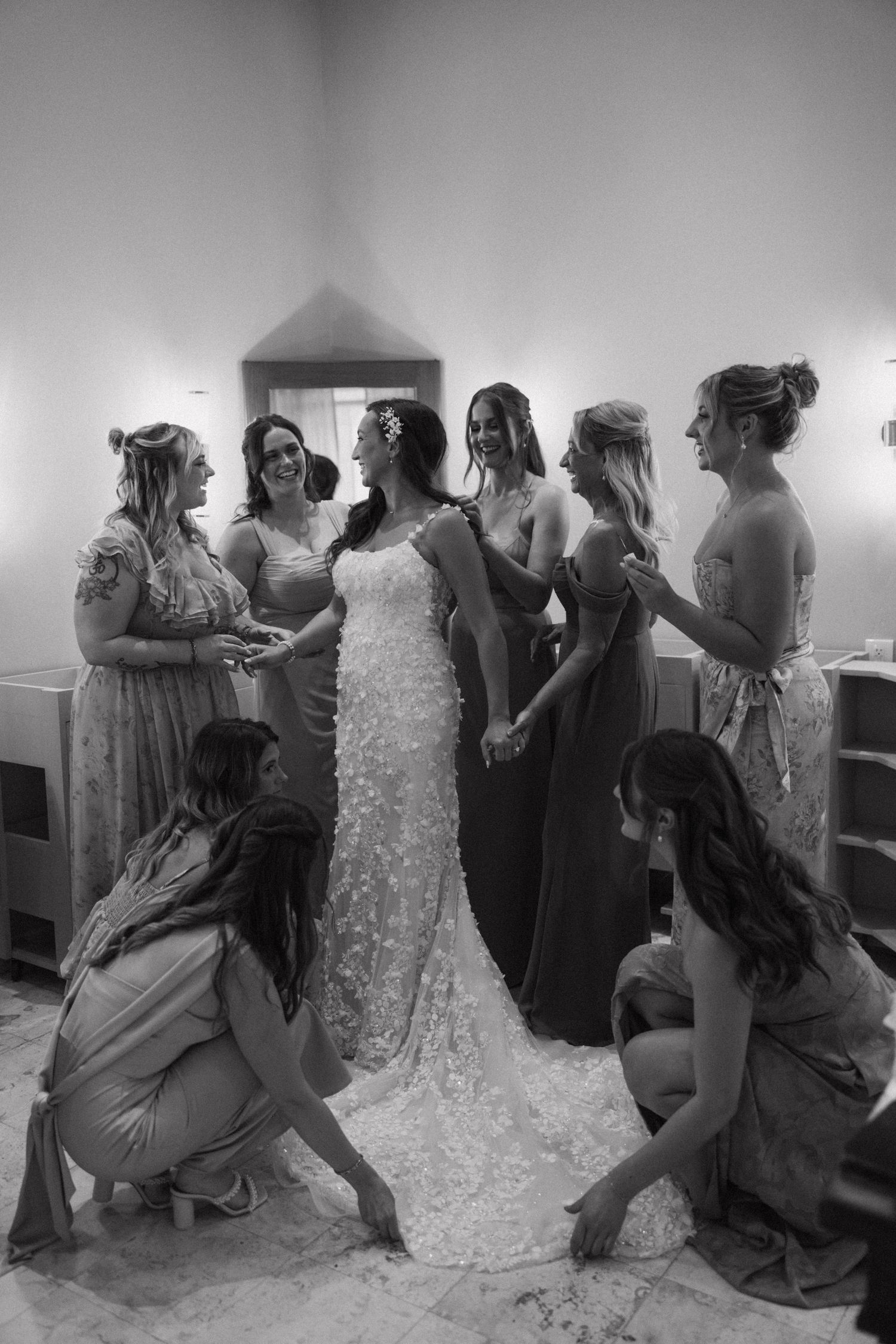 A group gathers around a wedding dress in a bridal preparation room.