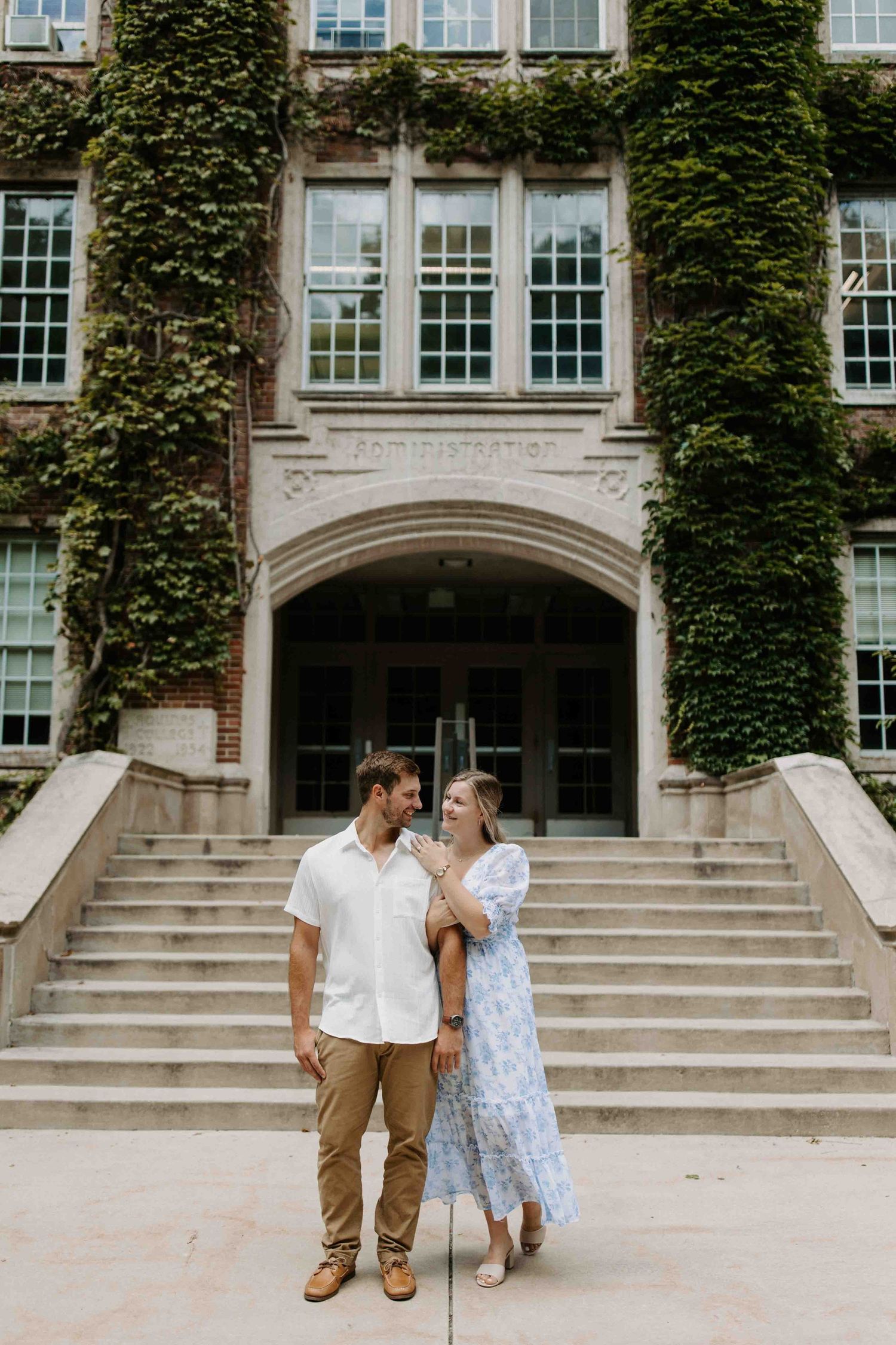 A couple shares a romantic moment on stone steps of an ivy-covered historic building.