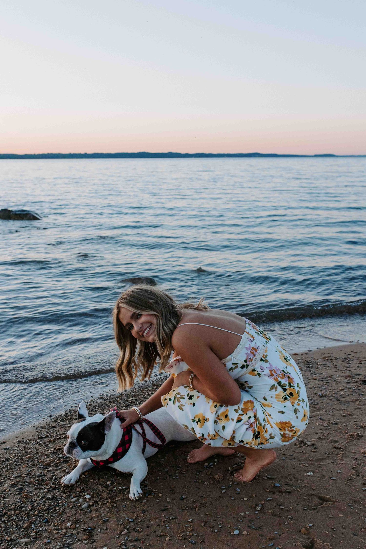 A person in a floral dress pets a black and white dog on a beach at sunset.
