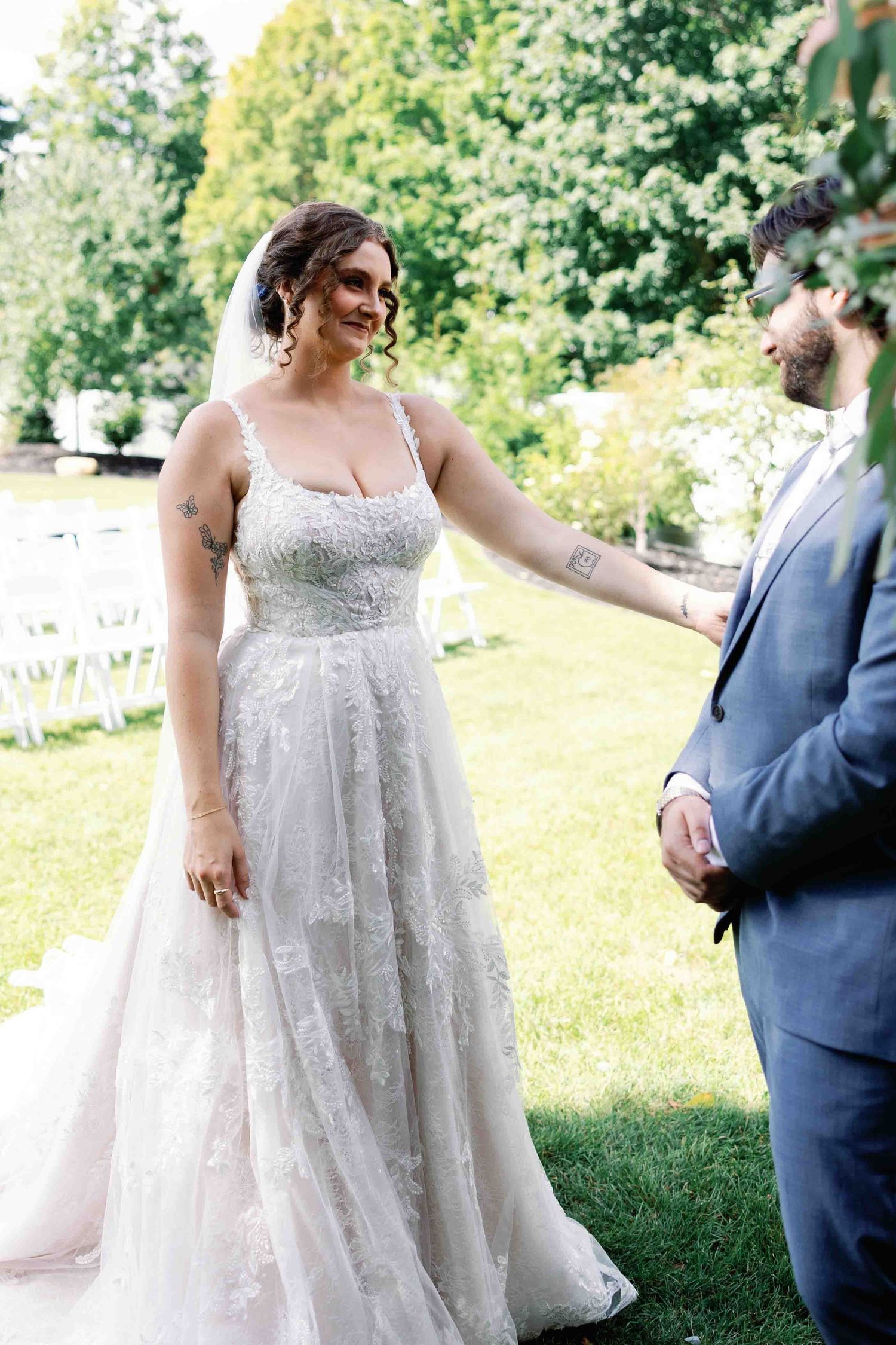 A bride in a beautiful lace wedding gown stands outdoors in a garden setting.