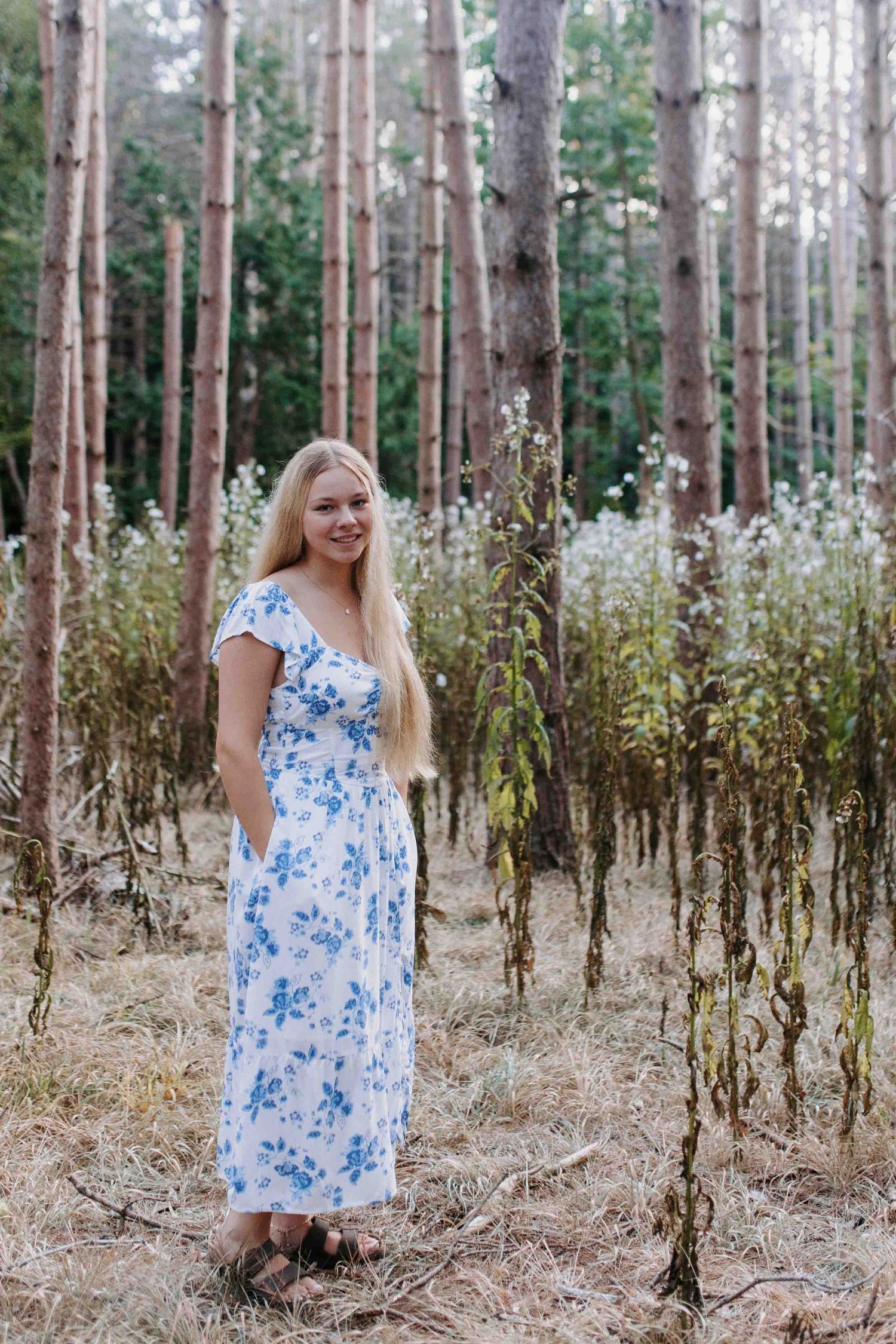 Person in blue floral dress smiles while standing in a pine forest.
