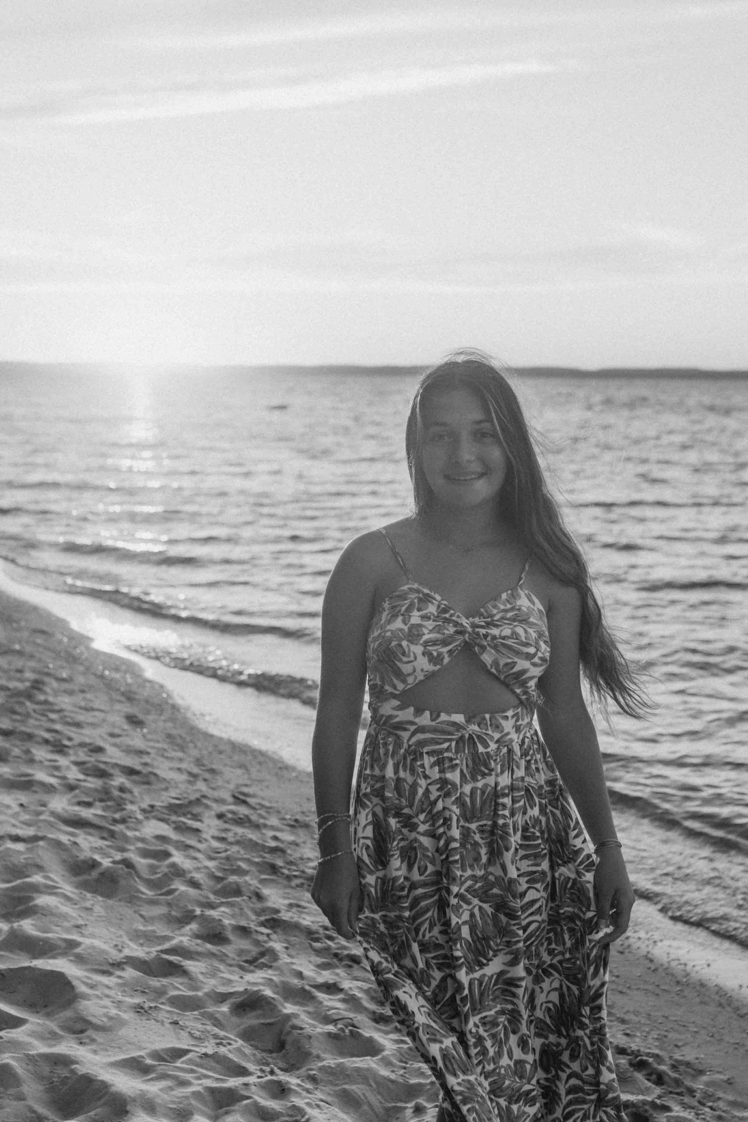 Black and white portrait of person in patterned dress on beach with ocean waves behind.