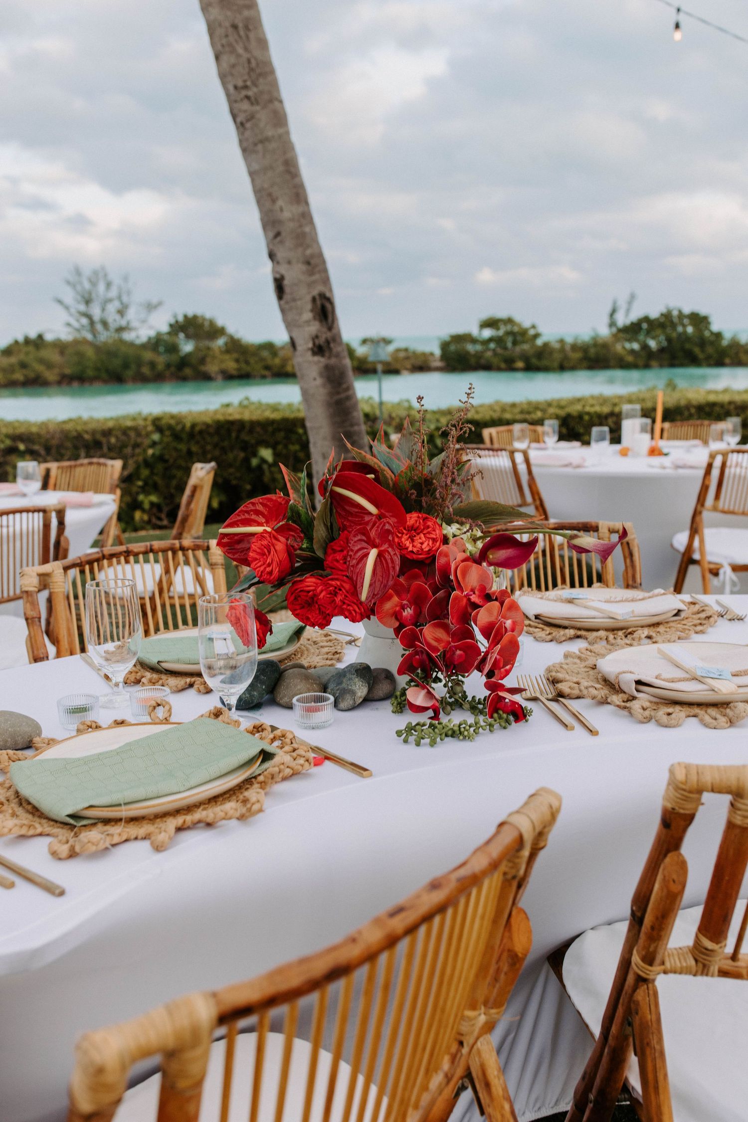 Elegant wedding table decor with red floral centerpieces and wooden chairs by the waterfront.