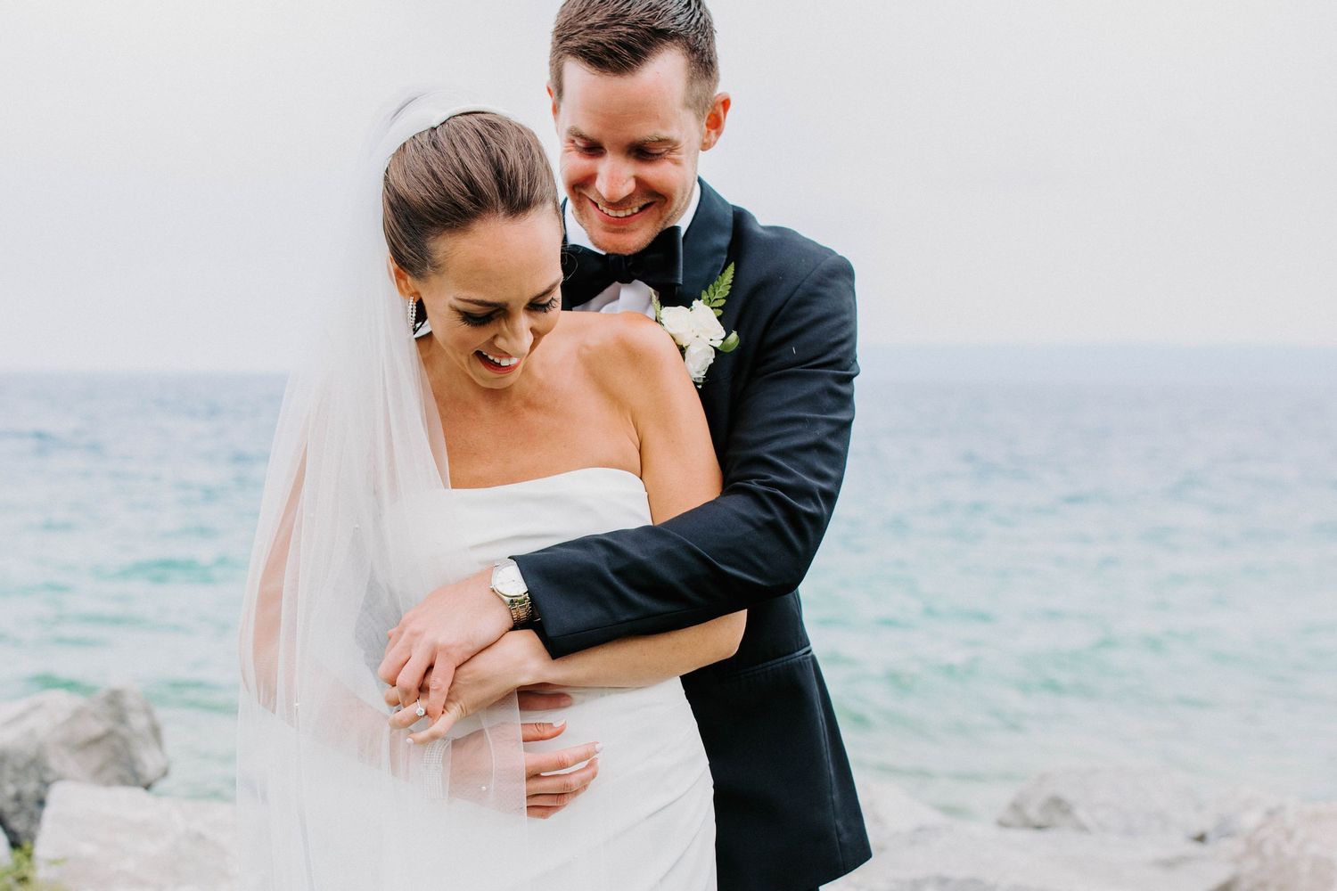 Candid wedding portrait showing couple laughing together by turquoise waters and rocky shore.