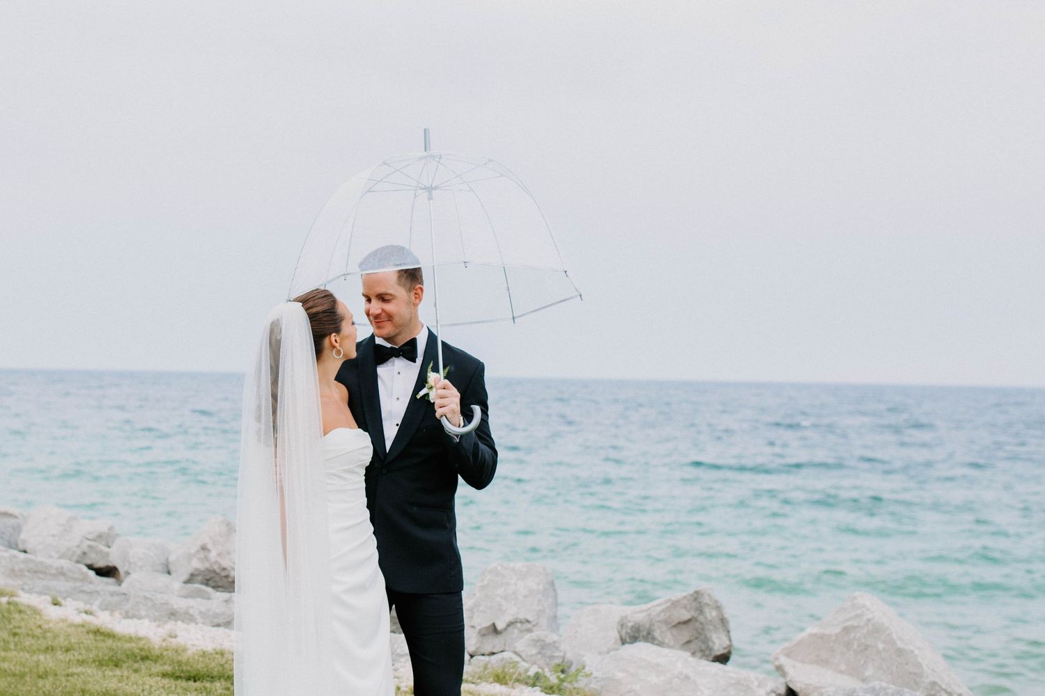 A seaside wedding portrait featuring a couple with a clear umbrella against a blue ocean backdrop.
